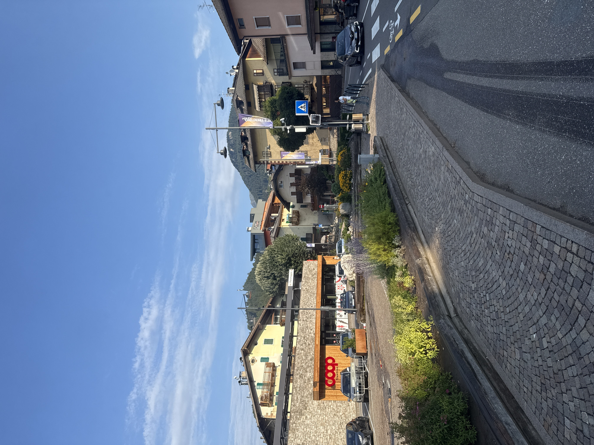 The image captures a street scene in Tesero, Italy, under a clear blue sky with scattered wispy clouds. In the foreground, a paved sidewalk made of small cobblestones runs alongside a road. To the left, a planter box with lush green and yellow foliage is visible.
Dominating the mid-ground is a building with a stone facade and a wooden-paneled entrance. The prominent "coop trentino" logo is displayed on the facade. Several cars are parked in front of the building. A streetlamp with two lights stands in front of the building.
Further back, several buildings with traditional alpine architecture, featuring yellow and pink facades, pitched roofs, and balconies, are visible. Mountainous terrain covered in trees forms the backdrop. On the right side of the road, a pedestrian crossing is marked with white stripes. A dark-colored car is crossing the pedestrian crossing, and a person is standing on the sidewalk near a street sign. Another person is visible walking on the sidewalk further down the street. The overall impression is of a bright, sunny day in a European town.