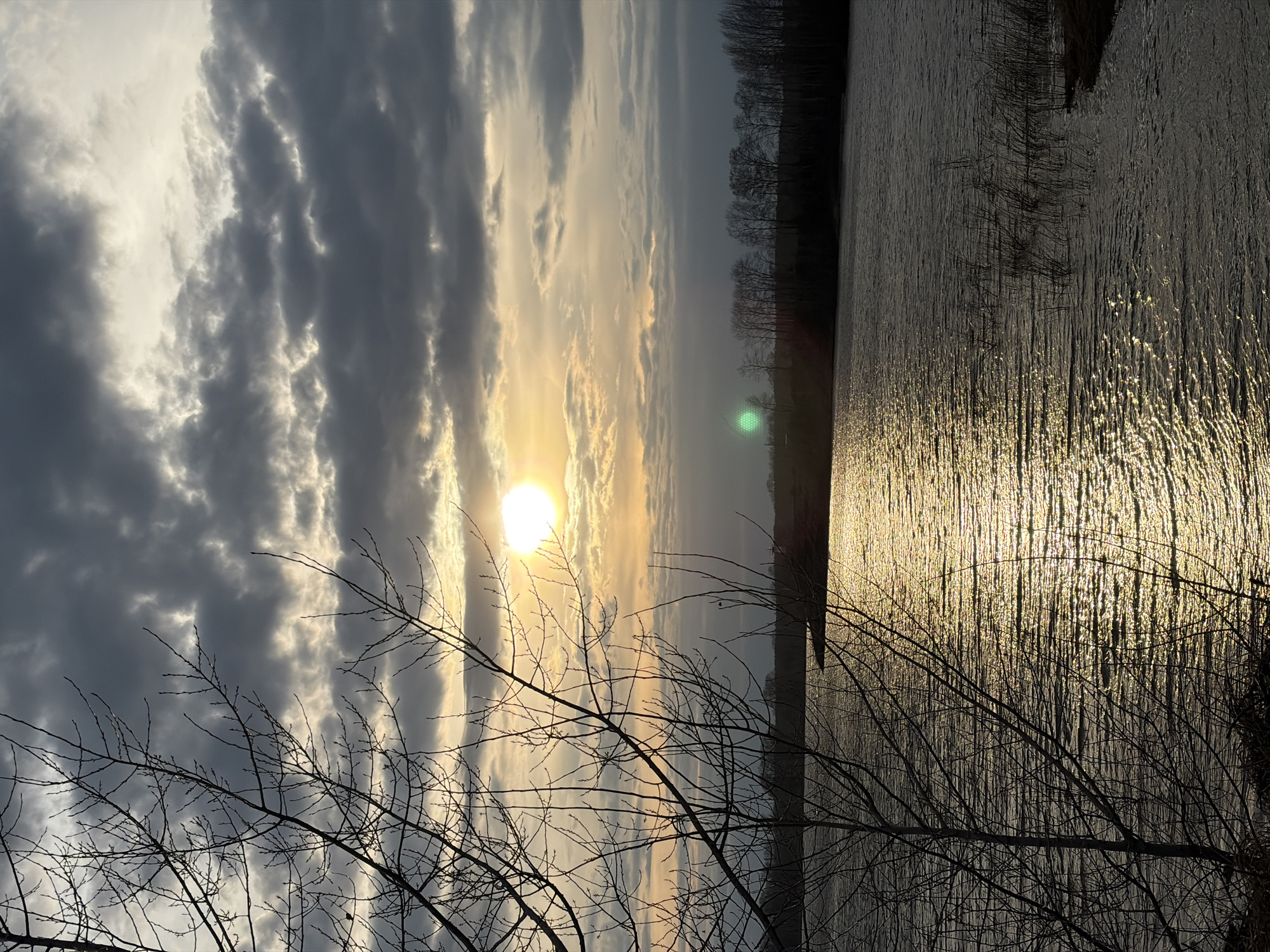 The image depicts a sunset over a body of water, likely a lake or reservoir, with tree branches silhouetted in the foreground. The sun is visible through a break in the clouds, creating a bright reflection on the water's surface. The sky is a mix of dark, cloudy areas and brighter, sunlit patches. A line of trees is visible on the far bank of the water. The location is likely in Chaikovsky, Russia, given the prompt's instructions.