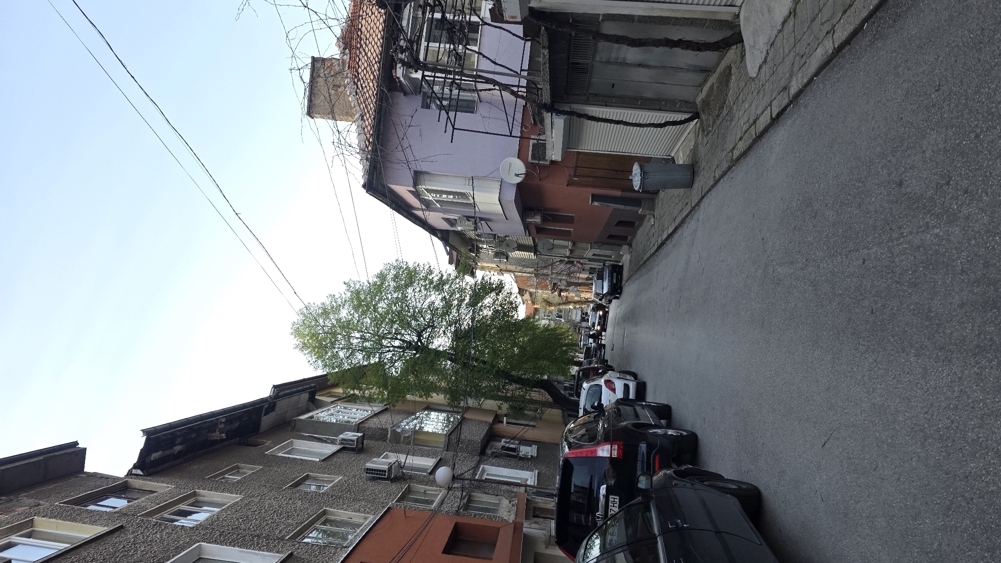 The image depicts a street scene in Blagoevgrad, Bulgaria. The road, constructed of asphalt, runs linearly into the background. Cars are parked along the right side of the road. Buildings flank both sides, varying in color and architectural style. A tree with green foliage stands between the buildings on the left. A trash receptacle sits near a building. Power lines traverse the sky overhead. The sky is clear and blue.