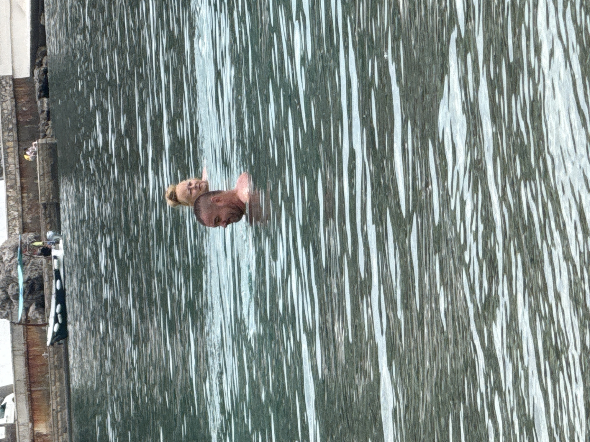 The image shows two people in the water near the coast, most likely in the city of Budva, Montenegro. A fair-skinned man with a shaved head is in the water with a fair-skinned woman with her hair up. The water has light streaks throughout. The coastline has a stone or concrete retaining wall. There are clothes or towels hanging on a line in the background.