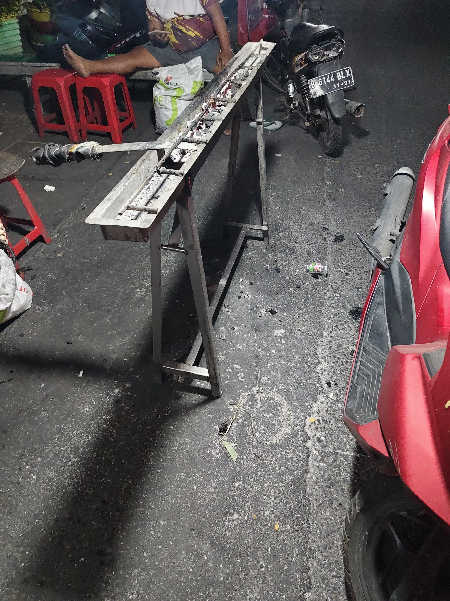 The image captures an outdoor scene, likely a street or sidewalk in North Jakarta, Indonesia. In the foreground, a red scooter is partially visible on the right side. To the left, a metal grill with embers and ash sits on a stand. A person's leg and foot are visible, resting on a red plastic stool. Beside the grill, a white bag with "CHEMICAL" printed on it is present. In the background, a black motorcycle with a license plate reading "B 6144 BLX" is parked. The ground is asphalt with scattered debris. The overall impression is that of a casual roadside setup, possibly related to street food preparation or sale.