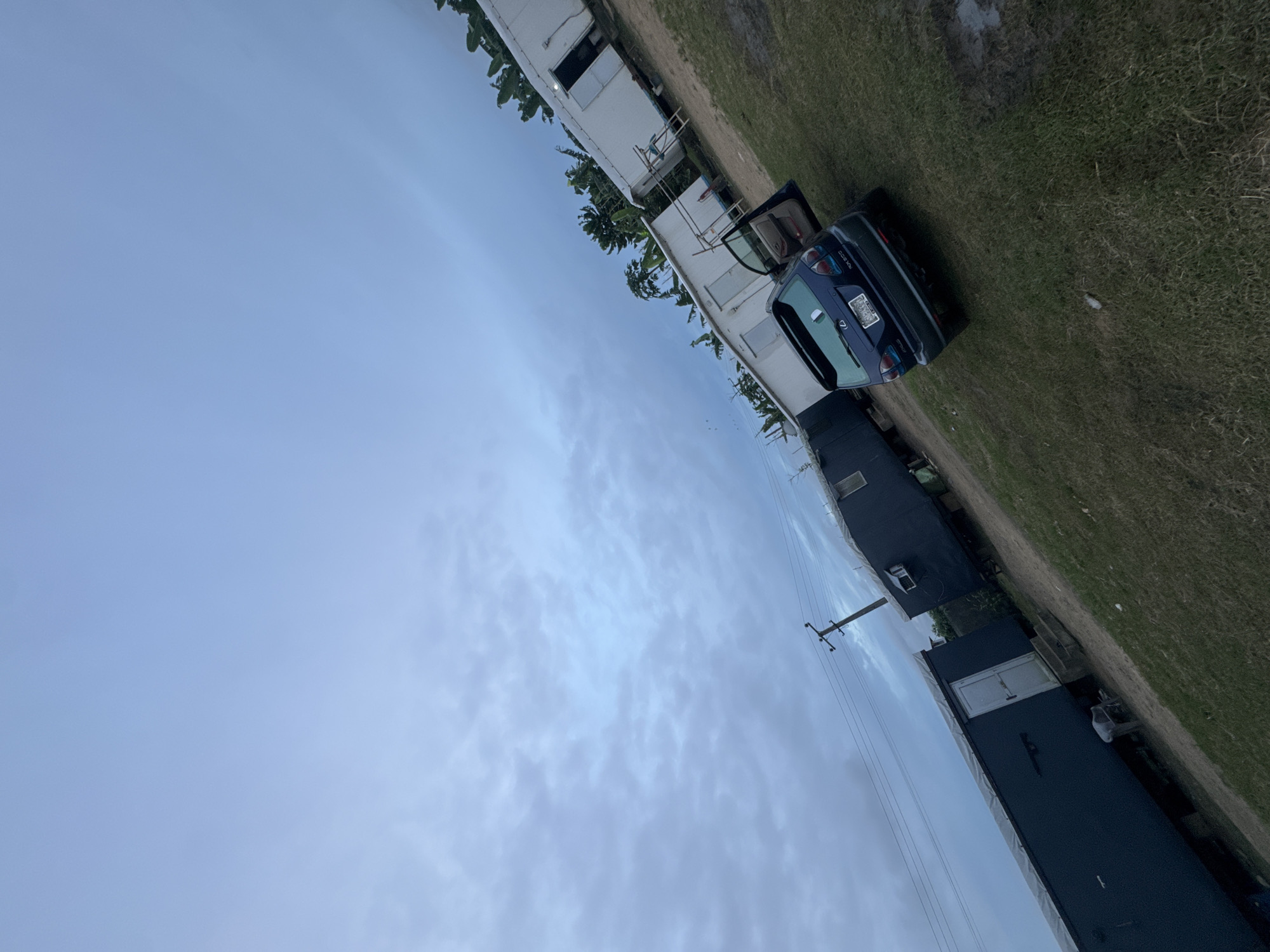 The image shows a scene in Igwuruta, Nigeria. A blue hatchback car with its driver-side door open is parked on a grassy verge next to a white mobile home or trailer. The surrounding environment includes more structures, potentially residential, some with dark roofs. A utility pole with power lines is visible. The sky is cloudy and pale blue. There are no people visible in the image.