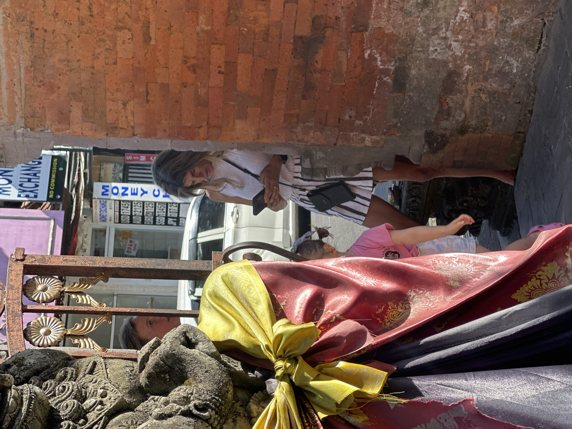 This media file shows a street scene in Ubud, Indonesia, likely during the daytime under bright sunlight. The foreground is dominated by ornate stone carvings and draped fabrics, primarily in shades of red and yellow, tied with a golden sash. A rustic metal gate with decorative elements is also visible.
In the mid-ground, a woman with long blonde hair, wearing a white sleeveless top and a striped skirt, is looking down at her phone, which she holds in her hand. She carries a black purse over her shoulder. Next to her, a young child wearing a pink shirt and white shorts is lying down, with their arm extended.
Behind them, a white vehicle is parked. A store facade is visible with signs indicating "MONEY EXCHANGE" and listing currency exchange rates for USD, AUD, EURO, SGD, and JPY. One sign also prominently displays "OPEN." The architecture includes brick walls, suggesting a somewhat rustic or traditional building style. The overall impression is a bustling street scene in a tourist-oriented location.