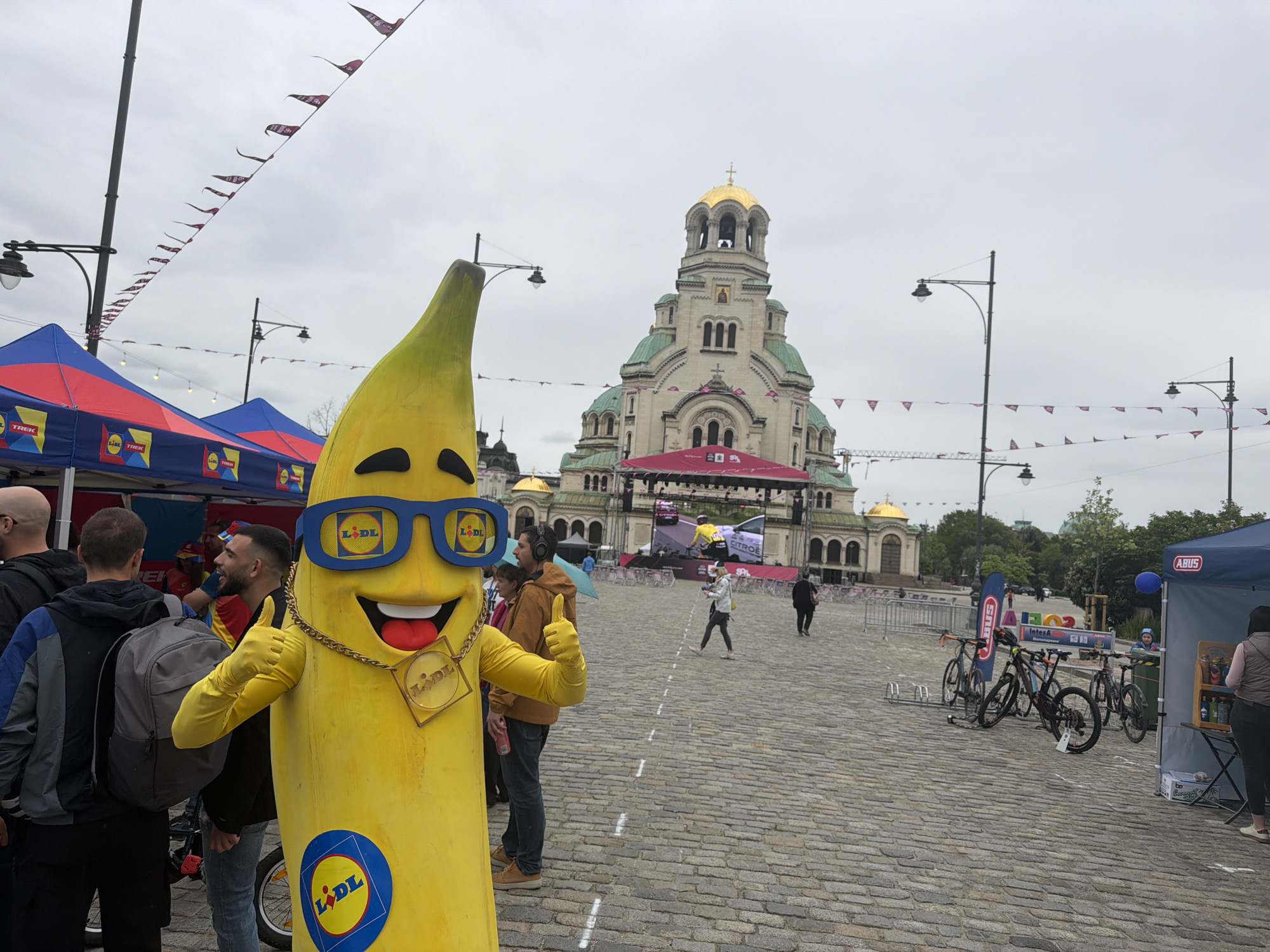 The image shows a large, yellow banana mascot, associated with the LIDL brand, standing in what appears to be a public square in Sofia, Bulgaria. In the background is the St. Alexander Nevsky Cathedral, a prominent landmark in the city. A series of flags are strung overhead. Several people are visible in the vicinity, along with tents, bicycles and related equipment. The ground is paved with cobblestones. A stage or large screen is set up in front of the cathedral.