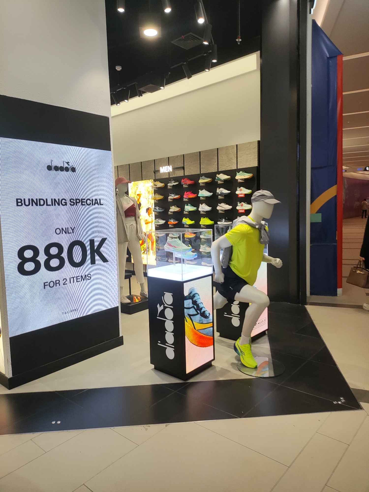 A retail store interior displays athletic footwear. On the left, a large digital screen advertises a "BUNDLING SPECIAL" for "880K FOR 2 ITEMS". To the right of the screen, a mannequin dressed in athletic wear, including a bright yellow t-shirt, black shorts, and neon green athletic shoes, is posed in a running stance. Behind the mannequin, shelves display rows of colorful athletic shoes. A second mannequin, wearing a light-colored tracksuit, stands further back in the store. The environment appears to be a modern shopping mall, with tiled flooring and suspended ceiling lighting. The store's branding, "diadora", is visible on the digital screen and a display stand. The scene is well-lit and appears to be an active retail environment. The location is specified as Lambangjaya, Indonesia.