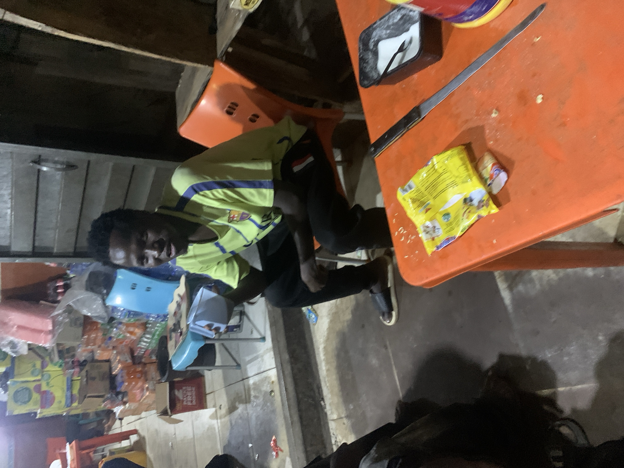 The image captures a dimly lit scene, likely indoors or semi-outdoors at night, possibly a shop or casual eatery in Gura Top, Nigeria. The main subject is a young man, seated at an orange plastic table, looking directly at the camera with a slight smile. He is wearing a light green and blue short-sleeved jersey, featuring an FC Barcelona crest on the left chest and what appears to be a Qatar Airways logo beneath it. A flag patch, with red, white, and black horizontal stripes, is visible on his right arm sleeve. He is also wearing dark pants and dark sandals.
On the orange table, a long, thin knife lies beside a yellow package of instant noodles, faintly identifiable as "INDOMIE Special Chicken." A small white container and a black tray with cutlery are also present on the table. The background is cluttered with shelves and various packaged goods, including bottles, boxes, and bags, suggesting a commercial setting. A blue plastic container or tray and a book are visible on a blue chair to the man's left. The floor appears to be concrete. A dark area in the bottom right foreground suggests the presence of another person or object.