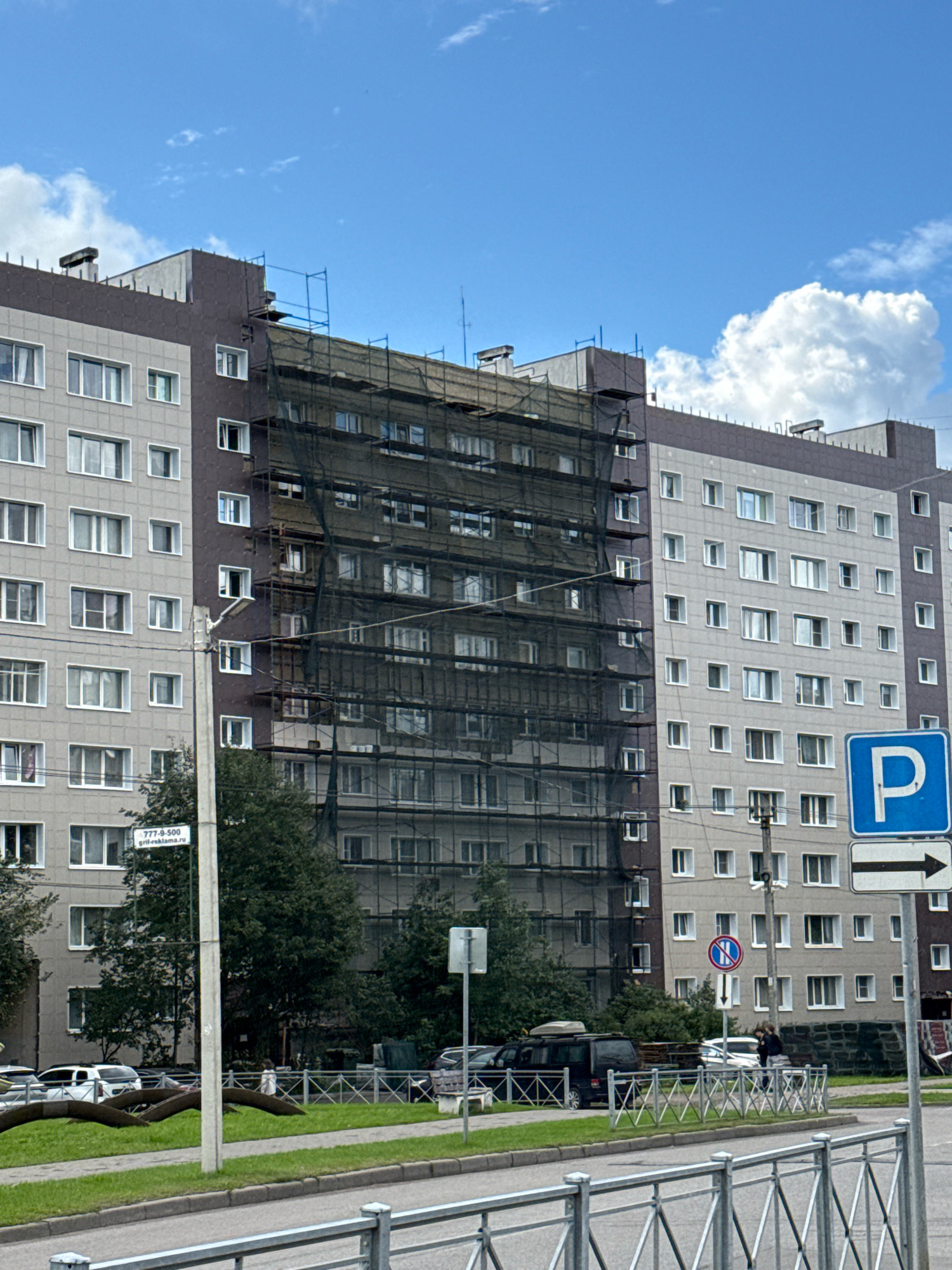 In the city of Vsevolozhsk, Russia, a multi-story apartment building is undergoing renovations. The building features a light gray facade on the left and a dark brown facade on the right. A significant portion of the building's facade is covered with dark netting, supported by scaffolding, suggesting exterior repair or insulation work. Several windows with white frames are visible on both sections of the building.
In the foreground, a street is visible with a gray asphalt surface and a metal railing. Beyond the railing, a sidewalk and a grassy area are present. Several trees are scattered throughout the scene, with some obscuring parts of the building.
A concrete lamppost stands near the building, with a sign attached to it displaying text and a phone number: "777-9-500" and "gril-reklama.ru".
Several vehicles are parked along the street. On the left, white cars are visible. In the middle, a dark minivan with a roof rack is parked. Further to the right, a white car is visible, and two individuals, one appearing to be an adult and the other a child, are walking near a fence.
In the background, blue and white traffic signs are mounted on poles. One sign is a blue square with a white "P" for parking, accompanied by a white arrow pointing to the right. Another sign below it is circular with a red border, depicting a horizontal blue bar with a red diagonal line through it, indicating "no stopping" or a similar restriction. A larger white sign with a blue circle and a diagonal red line is also visible, indicating a no parking zone.
The sky above is bright blue with scattered white clouds, suggesting a clear, daytime scene. The overall impression is a residential urban environment with ongoing construction work.