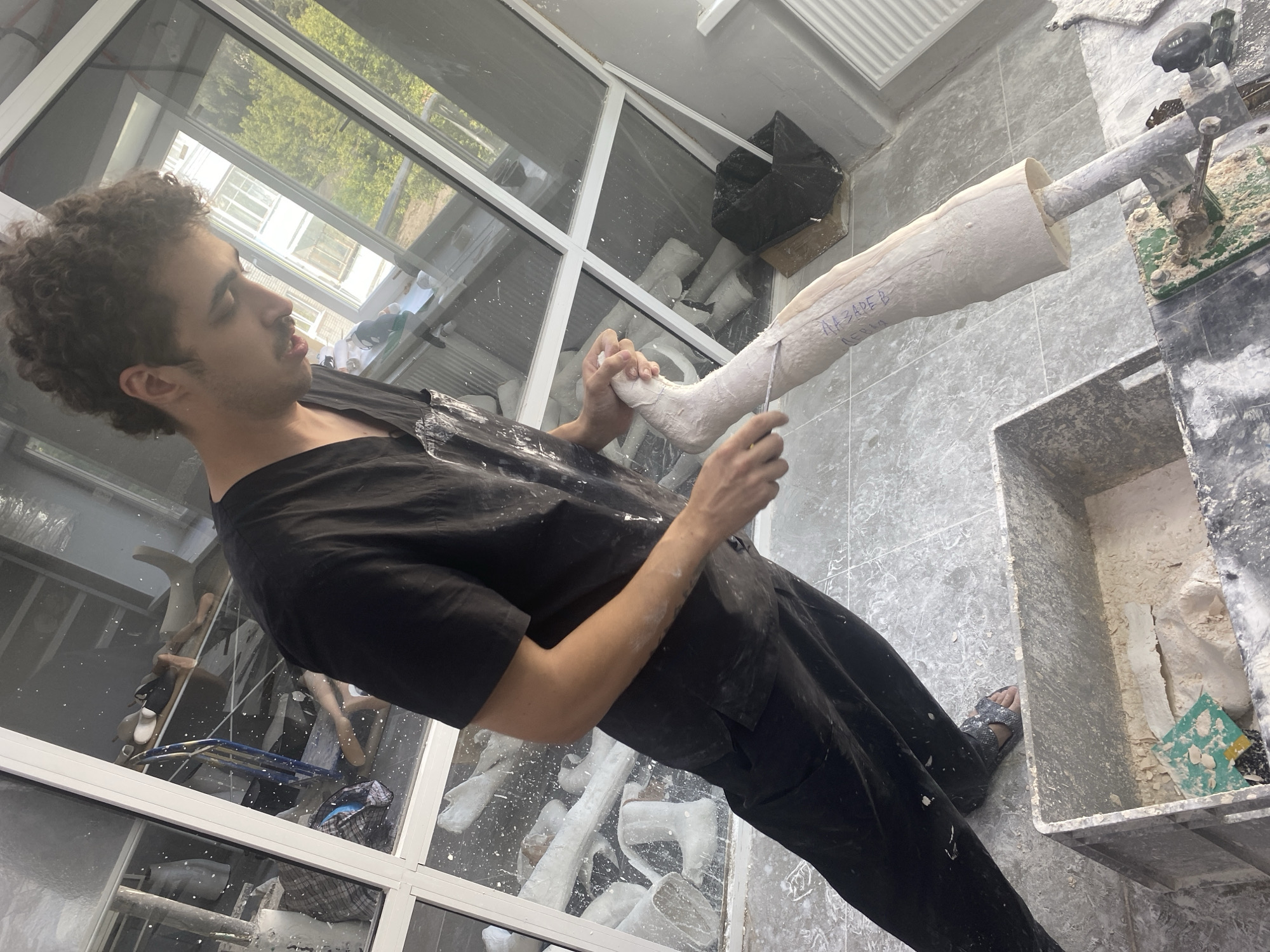 The image depicts a young man with curly dark hair, wearing a black t-shirt and black pants, engaged in a creative or technical process. He is holding a plaster cast of a limb, possibly an arm, with his hands. The cast is mounted on a metal rod that is secured by a clamp and a green platform. A white string is being manipulated along the cast. The setting appears to be a workshop or studio, with a floor covered in grey tiles. White plaster dust is visible on the floor, the man's arms, and various surfaces, suggesting an ongoing project.
In the background, large windows reveal a glimpse of the outdoors with trees and buildings, indicating it is daytime. Through the windows, other plaster casts and studio equipment can be seen, including what appear to be mannequins and stands. There is a large black bin and a grey plastic container filled with plaster debris and tools. The overall atmosphere suggests a focused and hands-on activity. Some text is visible on the plaster cast in blue marker, reading "НАЗАРЕВ В" and below it "ПЕРВАЯ". The location is identified as Vladimir, Russia. The man's expression is concentrated, indicating his engagement with the task.