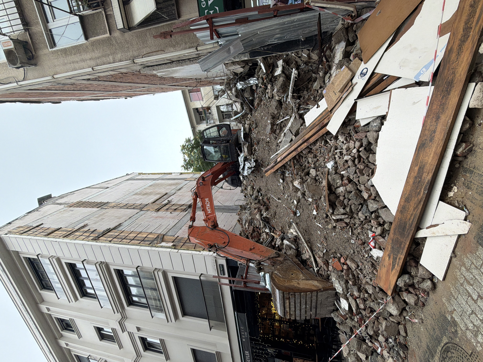 The image depicts a construction site in Kadıköy, Turkey, characterized by debris and heavy machinery. An orange Hitachi excavator is positioned atop a large pile of rubble, which includes bricks, concrete, and wood. To the left of the excavator, a modern building with balconies and large windows stands in contrast to the surrounding demolition. A shop with a "MUSIC STORE" sign is visible at the base of this building. To the right, an older, weathered building with visible brickwork and an air conditioning unit is present. Construction fencing and warning tape are present, indicating a work zone. The sky is overcast. No people are clearly visible in the image.