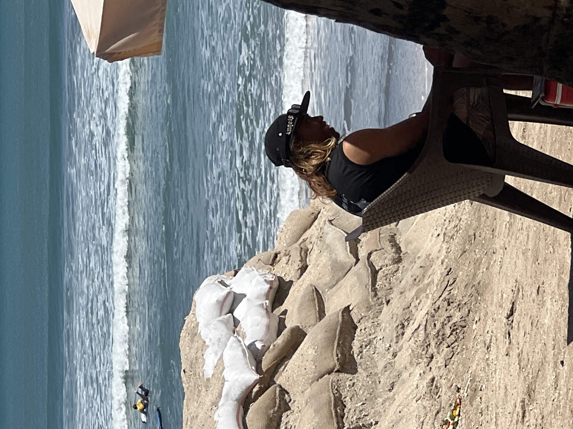A person sits in a chair on a sandy beach, facing the ocean. The person is wearing a black t-shirt and a black baseball cap. They have long, light brown hair. In the background, waves are breaking on the shore. There are sandbags piled up on the left side of the image, and a beach umbrella is visible on the right. In the distance, two people are in the water on surfboards. The location is Kuta Beach, Bali, Indonesia.