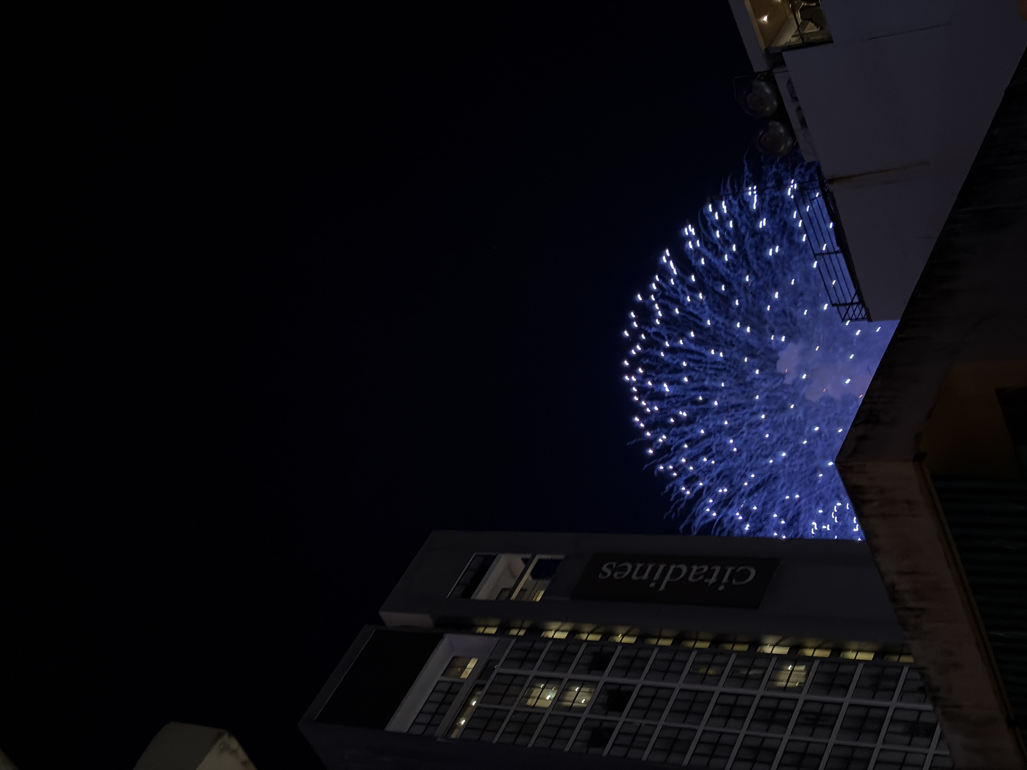 A nighttime outdoor shot captures a wide view of a building in Nha Trang, Vietnam, as fireworks burst in the sky. The building, partially illuminated by internal lights, has a dark facade with numerous windows. A banner with the word "citadines" is visible vertically on the side of the building. In the background, a large, bright blue fireworks display explodes in a circular pattern, with streaks of light illuminating the dark sky. The foreground shows the edges of other buildings, one with a stone-like texture on its upper surface.