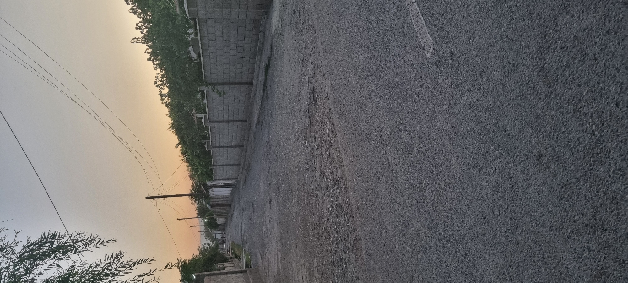 The image depicts a street scene in Havaskor, Tajikistan, during what appears to be dusk or dawn. The road surface is paved with asphalt, showing some signs of wear and tear with visible potholes and unevenness. On the right side of the road, a long, grey brick wall runs the length of the image, topped with leafy green trees and vines. On the left side, a similar brick wall is visible at the beginning of the scene, with more trees and vegetation extending further down the road. Electrical wires and poles are prominent in the background, stretching across the pale, softly colored sky, which transitions from a light grey at the top to a faint orange hue near the horizon. A few indistinct figures of people can be seen in the distance on the left side of the road, walking away from the camera. The overall atmosphere is quiet and tranquil, characteristic of a residential area at the end or beginning of the day.
