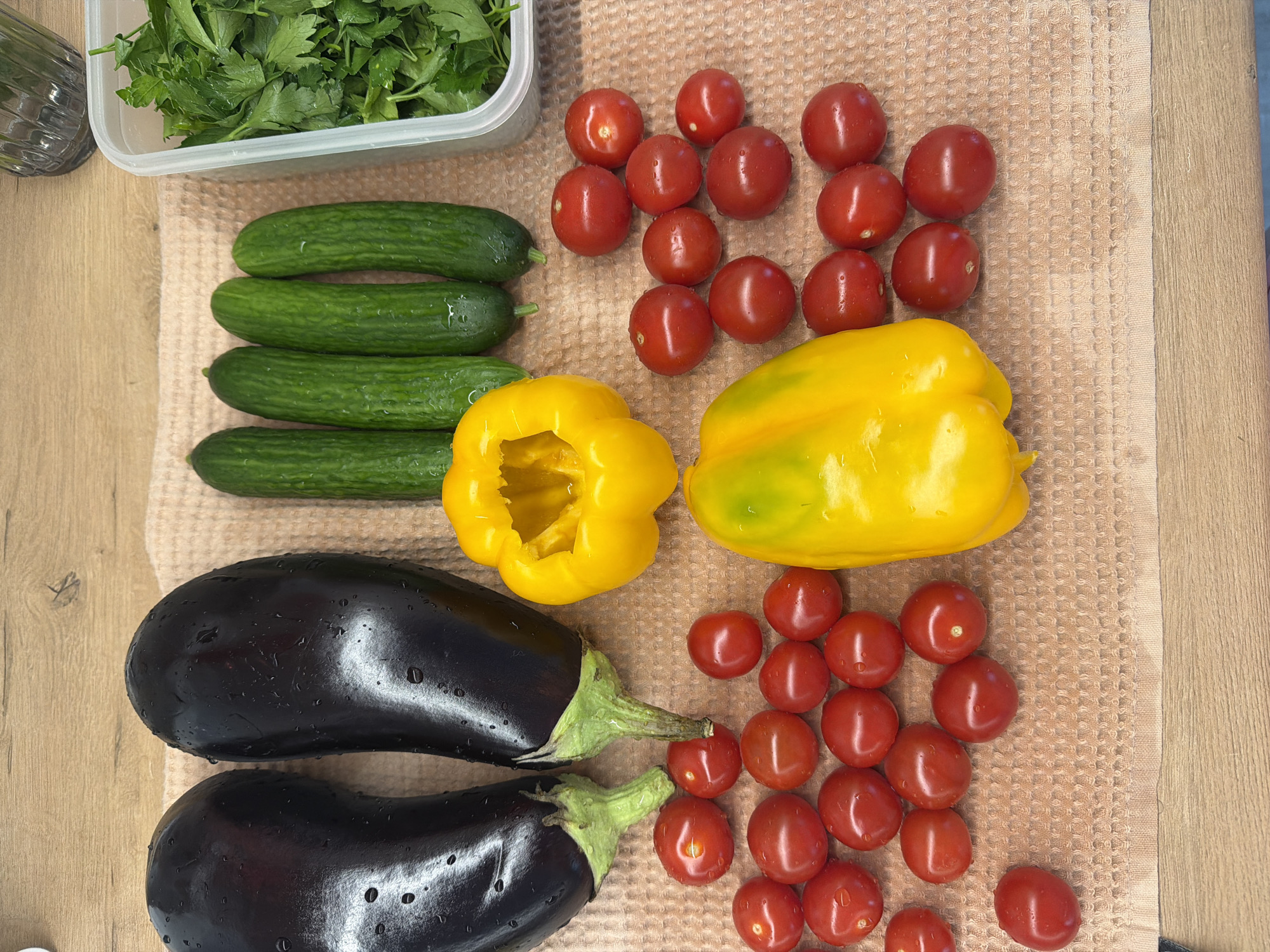 A high-angle shot depicts an arrangement of fresh vegetables on a light brown textured towel, possibly a kitchen towel, placed on a wooden surface with a visible wood grain.
The vegetables include:
* Four small cucumbers, grouped together at the upper left of the frame.
* Two dark purple eggplants, positioned at the bottom left.
* Two yellow bell peppers, one with the top cut off, centrally located.
* Approximately 25 red cherry tomatoes, clustered in two groups, one at the top and one at the bottom right of the frame.
* A clear rectangular container with green leafy herbs at the top left corner.
The vegetables appear fresh and clean, with water droplets visible on some surfaces.
The background is blurred, suggesting a shallow depth of field. There is no evidence in the image to indicate the location is Votkinsk, Russia. No people, actions or interactions are discernible from the image.