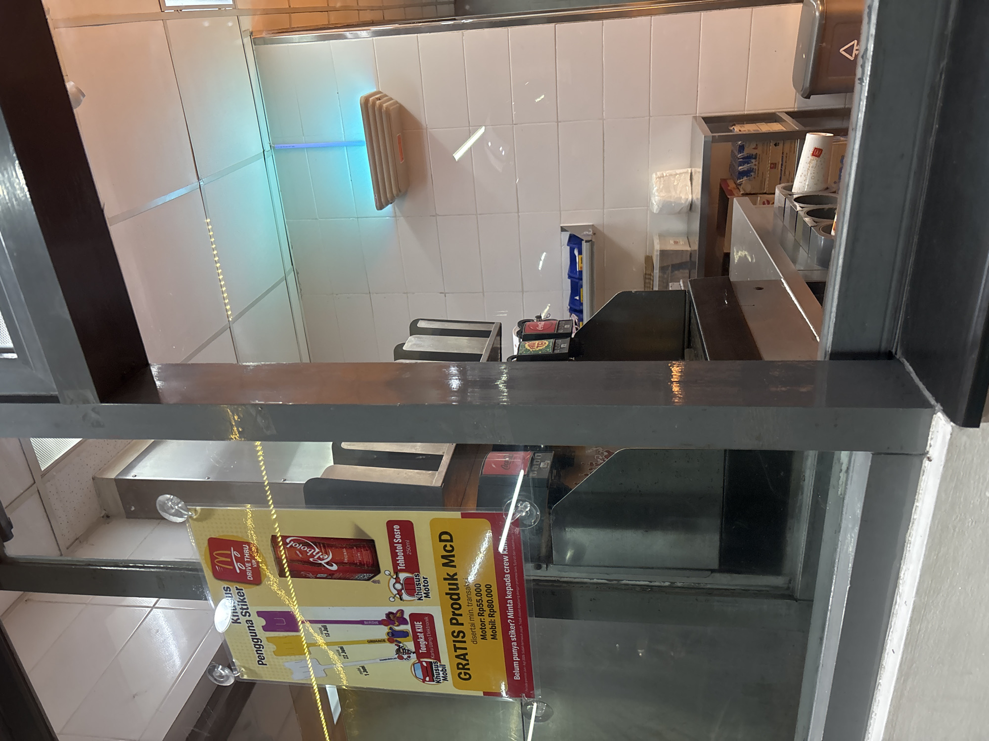 This image, rotated on its side, provides a clear view through a service window, likely a drive-thru, into a McDonald's fast-food preparation area in South Tangerang, Indonesia. No people are present in the scene.
The primary subject is a promotional sign, held by suction cups onto the glass window, facing the viewer. It features the McDonald's golden arches logo and the text "DRIVE THRU VIP". The sign announces "GRATIS Produk McD" (FREE McD Product) for "Khusus Pengguna Stiker" (Special for Sticker Users), with minimum transaction requirements of "Rp55.000" for motorcycles ("Motor") and "Rp80.000" for cars ("Mobil"). It also encourages customers to "Minta kepada crew kami" (Ask our crew) if they don't have a sticker. The promotion includes "Tehbotol Sosro 250ml" and a collectible "Tongkat KUE" (cake stick) associated with electronic money card transactions, featuring characters like Birdie, Grimace, and Hamburglar. Partially visible dates such as "13 Juli", "22 Juli", and "1 Agustus" indicate the promotional period.
Behind the window, the service area features a dark counter with various items including drink cups (some with the McDonald's logo), a drink dispenser, and other equipment. The background wall is covered in white tiles, and a prominent light fixture emits a striking blue glow. Stacks of supplies and boxes are visible on shelves and counters further back. The overall lighting is artificial and bright, typical of a commercial establishment.