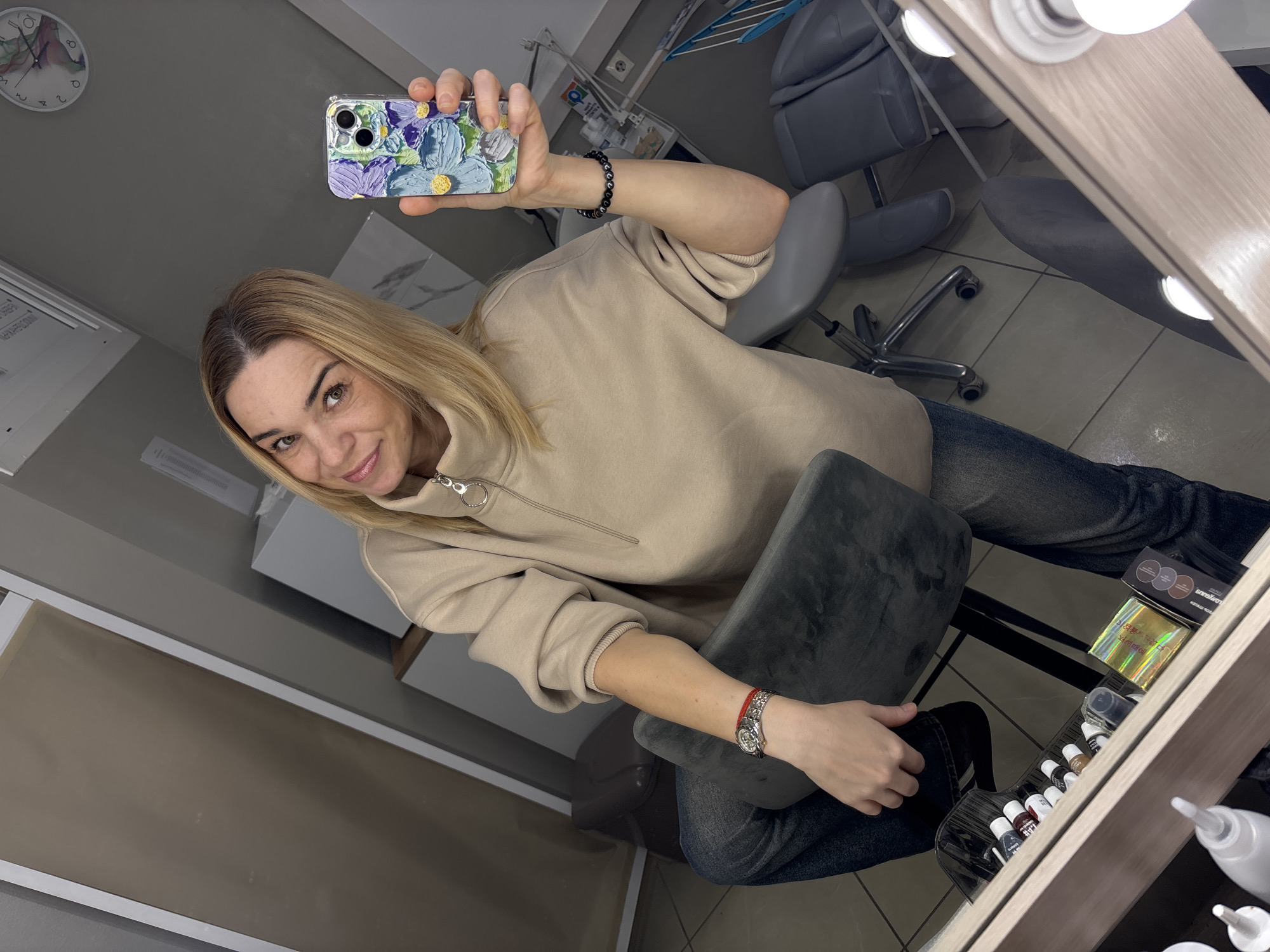 A woman with blonde hair is taking a selfie in a room with a mirror. She is wearing a light brown hoodie and blue jeans. On her left wrist, she has a watch with a silver band and a red bracelet. Her phone, which has a floral case with purple and blue flowers, is visible in her right hand. The room appears to be a salon or a beauty studio, with chairs, tables, and shelves of products, including small bottles and containers of what might be makeup or nail polish, reflected in the mirror. There are also lights above the mirror, suggesting it's a vanity area. A clock on the wall shows it is approximately 10:50. The woman is smiling and looking at the camera. The overall mood seems casual and self-assured.