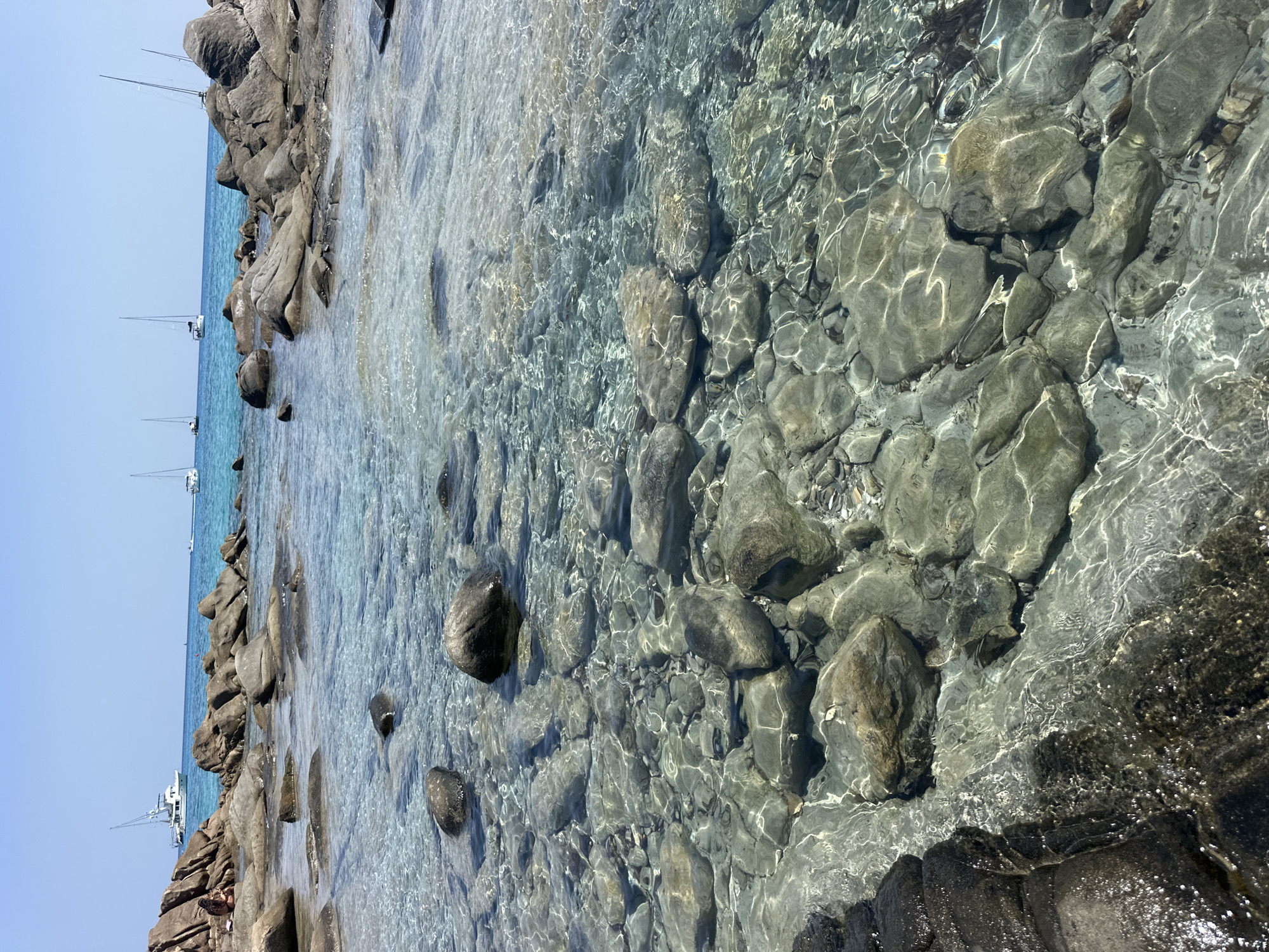 The image depicts a sun-drenched coastal scene in Crabonaxa, Villasimius, Italy. The foreground is dominated by clear, shallow turquoise water revealing a rocky seabed. Numerous smooth, grey and brown stones of varying sizes are submerged, with sunlight creating rippling patterns on their surfaces. In the mid-ground, larger, rougher rocks emerge from the water, forming a natural barrier.
Beyond the rocks, the sea transitions to a deeper blue, and several sailboats are visible on the horizon, their white sails stark against the clear blue sky. One person, appearing as a small figure, sits on the rocks on the far left side of the image, facing away from the camera and towards the sea. The overall impression is one of a tranquil, picturesque Mediterranean beach.