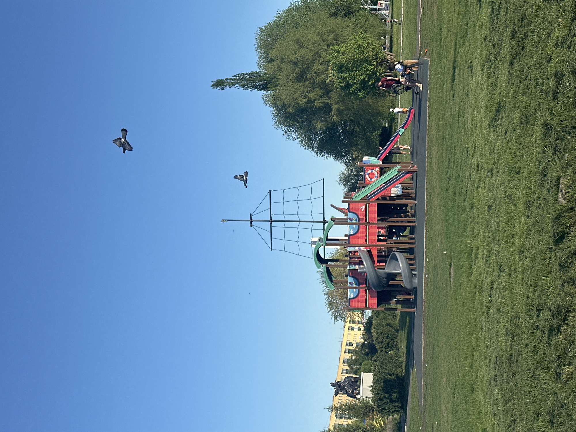 The image shows a playground in a park, set against a clear blue sky in Unknown, Unknown. The playground features a large, colorful pirate ship structure with climbing nets, slides, and various play elements. The playground is surrounded by grassy areas, with trees and a statue visible in the background. Two birds are flying in the clear sky above the playground. In the background, there's a building with multiple windows, and further back, additional park visitors can be seen.