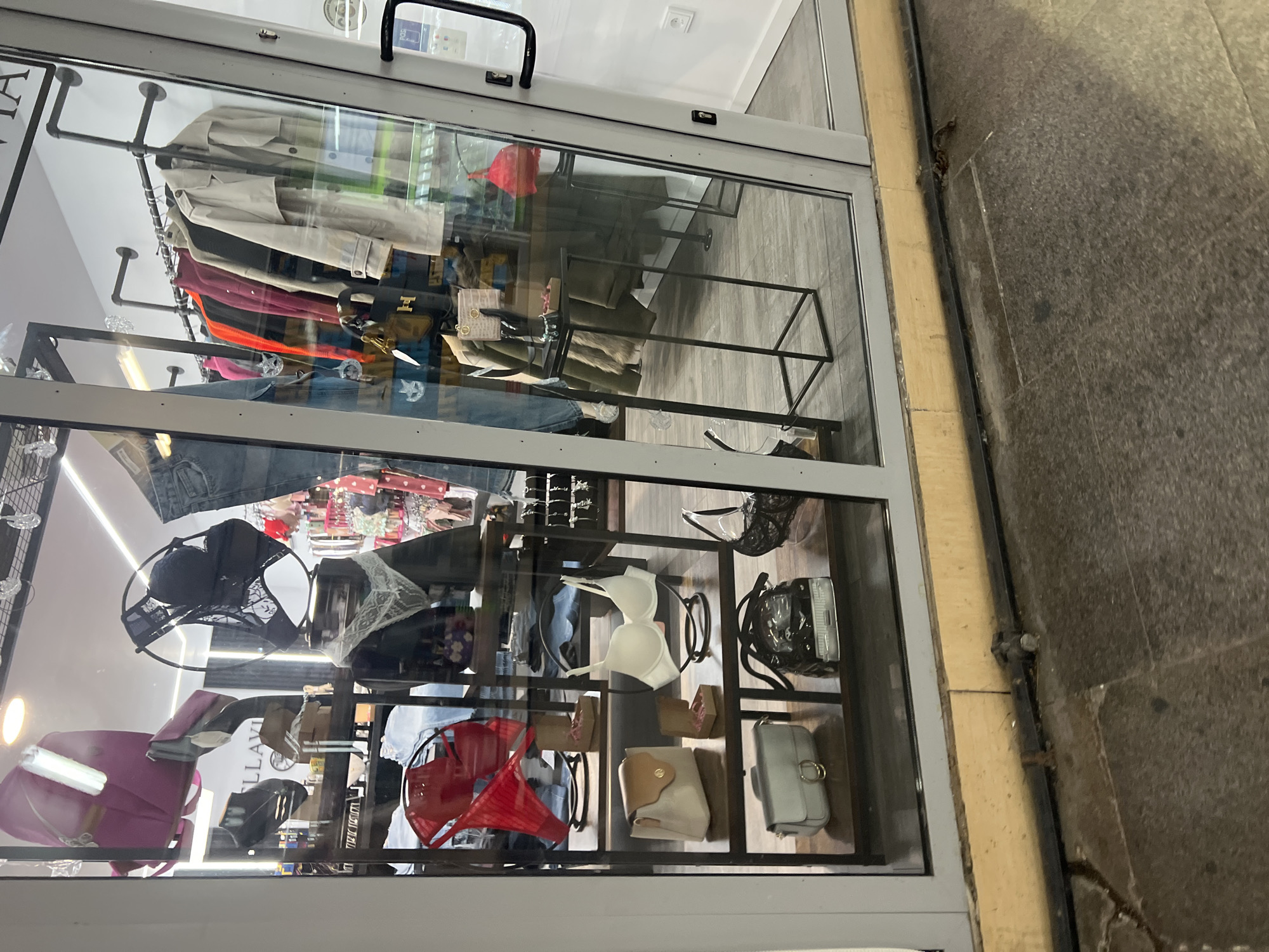 The image captures the window display of a clothing store in Plovdiv, Bulgaria. The window is filled with a variety of clothing items, including dresses, blouses, skirts, jeans, and bags. The clothing is displayed on shelves and mannequins. The store appears to be located on a busy street. The sidewalk outside the store is made of stone.