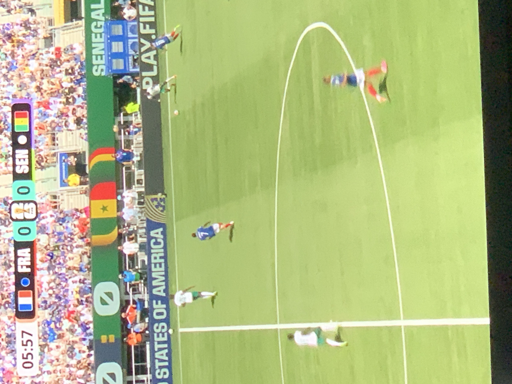The image depicts a soccer match at a stadium, with a digital scoreboard displaying "05:57" for the time and "FRA 0 0 SEN" for the score, indicating France and Senegal are playing. The scoreboard also shows FIFA logos. The field is green with white lines. In the foreground, several soccer players are visible on the field. One player in a white uniform is running towards the left side of the frame, and another player in a blue uniform with the number 7 is running towards the right. In the background, a large crowd is seated in the stadium stands, and flags are visible. The word "SENEGAL" is displayed on a banner in the background. The phrase "PLAY.FIFA" is also visible on a dark banner. The scene appears to be an outdoor event. The question asks for the location in the city of Barcelona, Spain, but there is no visual information in the image to confirm or deny this location.