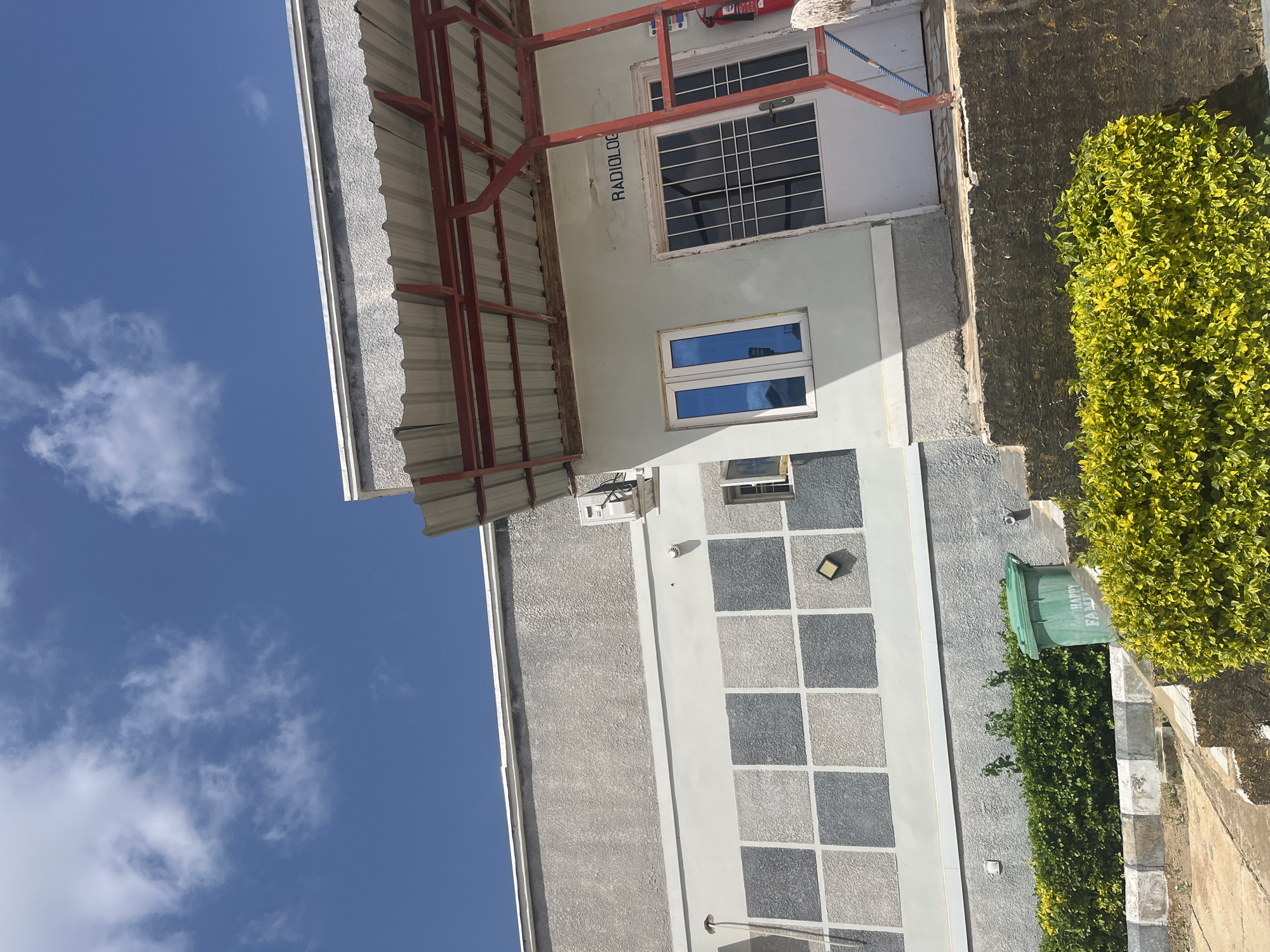 The image shows the exterior of a building with the word "RADIOLOGY" visible above a doorway. The building appears to be a medical facility. In the foreground, there are steps leading up to the building and lush green and yellow bushes. A green trash can is situated near the steps. The sky is blue with scattered white clouds. The building has a light-colored facade with a patterned section and windows. There is also an awning or overhang with a red metal frame. The scene is set in the city of Jos, Nigeria.