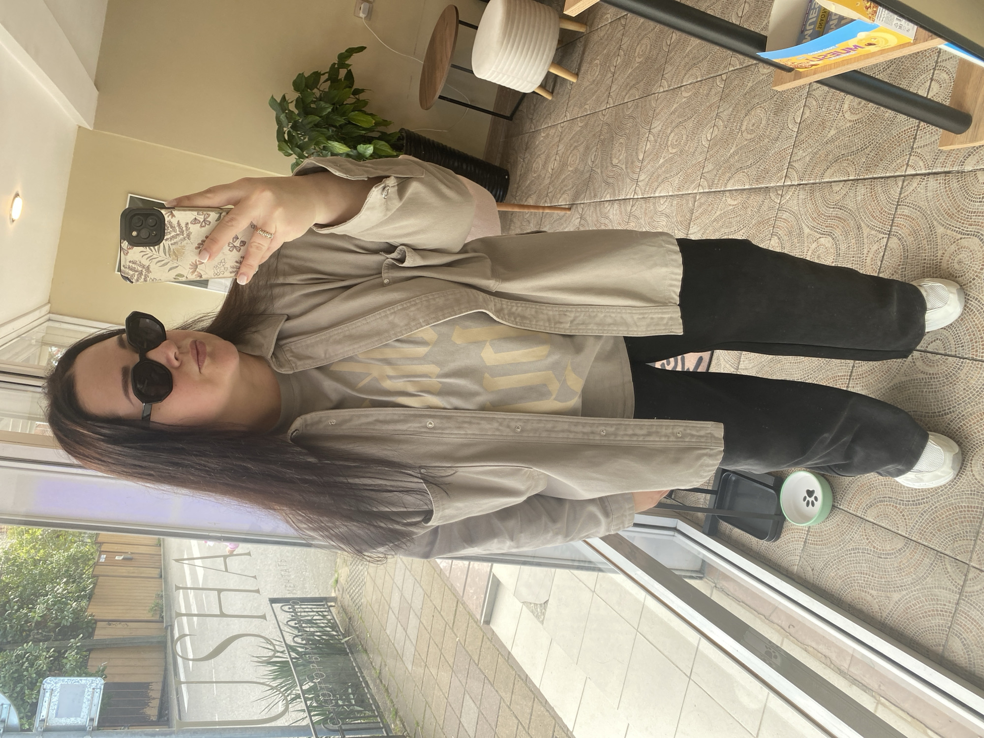 A person wearing sunglasses, a beige jacket, a light gray t-shirt, and black pants is standing near a window. They are holding a smartphone with a patterned case, presumably taking a photo or video. Outside the window, there is a view of a street with trees and buildings. A sign in Russian is partially visible on the window itself. Inside, there is a stool with a white padded top, a plant in a black pot, a magazine rack, and a patterned tile floor. A pet bowl is located near the feet of the person. The text in Russian on the window reflects the letters "Ю.C.H.A" which can be transliterated to "Yu.S.H.A" (ЮША), indicating a location that may be related to the name "Юша" in Sochi, Russia.