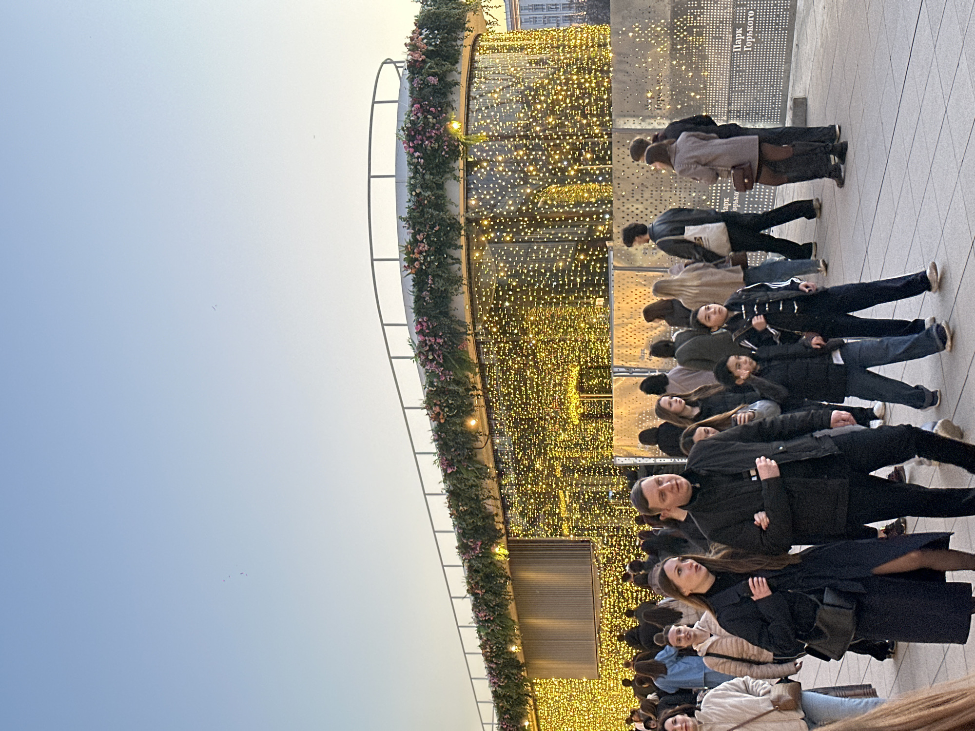 A large group of people of diverse ages stand and walk on a paved surface. Behind the group, a structure with a decorative arrangement of lights is visible. Part of the structure is adorned with greenery and flowers. The background also includes buildings and a sign with the words "Парк Горького" or "Park Gorkogo" indicating that this scene is most likely taken in Moscow, Russia. The sky is visible at the top of the image, with a gradient of light colors.