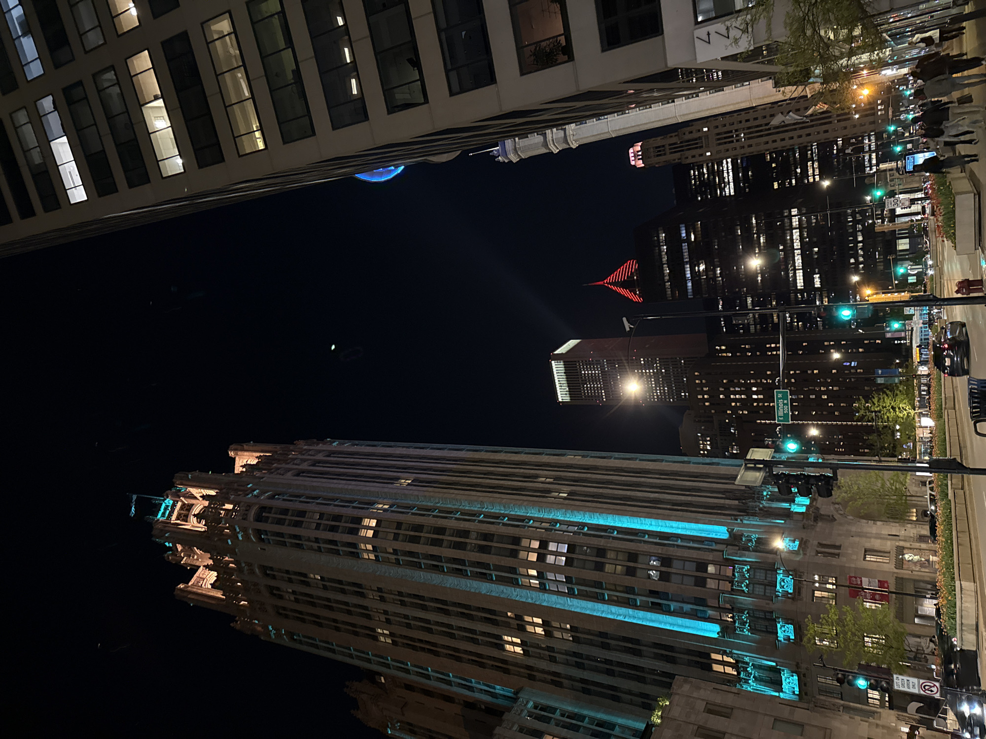 The image is a nighttime view in Chicago, Illinois, United States of America, taken from a low angle looking upwards at several buildings. The building on the left side is a skyscraper with many windows, some of which are illuminated. Above, the Blue Cross Blue Shield building has a red neon logo at the top.
On the right side of the image is a portion of a city street with sidewalks, traffic lights, trees, and pedestrians walking. A street sign visible reads "E Illinois St 500 W".
The building on the lower right has decorative architectural elements and is illuminated with a blue-green light.