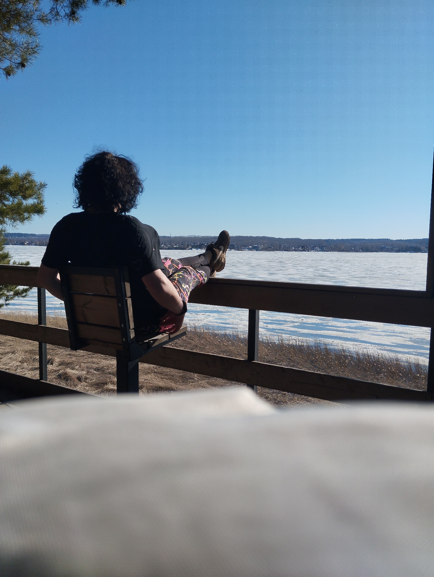 The image shows a person sitting on a bench, facing a frozen lake. The person has dark, curly hair and is wearing a black t-shirt and colorful leggings with black shoes. Their feet are resting on a wooden railing that surrounds the bench. In the background, a body of water mostly covered in ice stretches toward a distant shoreline with a horizon of land and buildings. The sky is clear and blue. The location could be in a park area overlooking a water body in Votkinsk, Russia, but this is speculative.