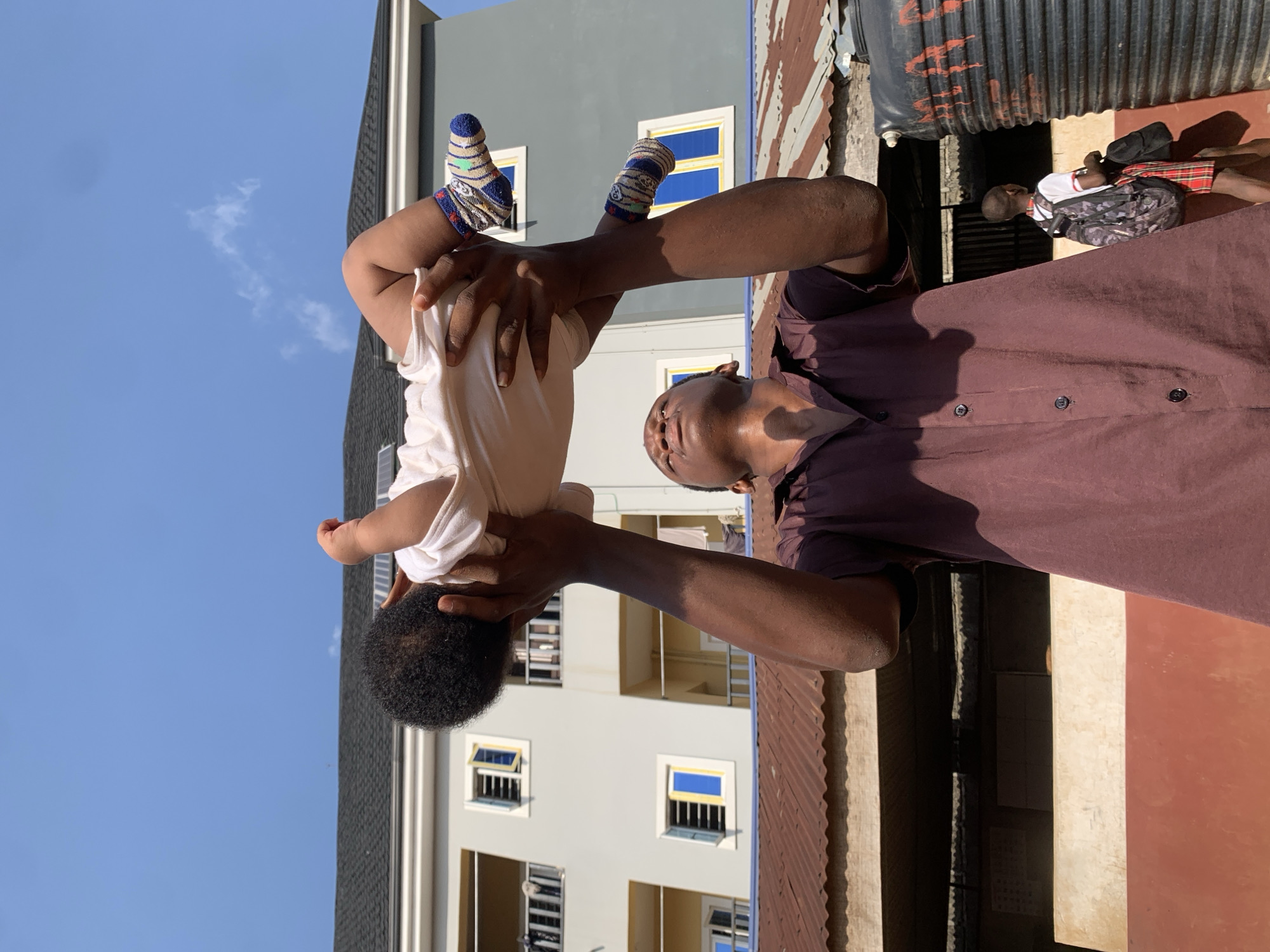 In Ukwu-Abwa, Nigeria, an adult holds a baby upside down, with the baby's head pointing downward. The adult wears a buttoned brown shirt. The baby wears a white shirt and socks with blue and yellow stripes. Behind them is a light-colored building with blue-trimmed windows and a blue sky. Another individual stands behind the adult, obscured by shadow and a water tank.