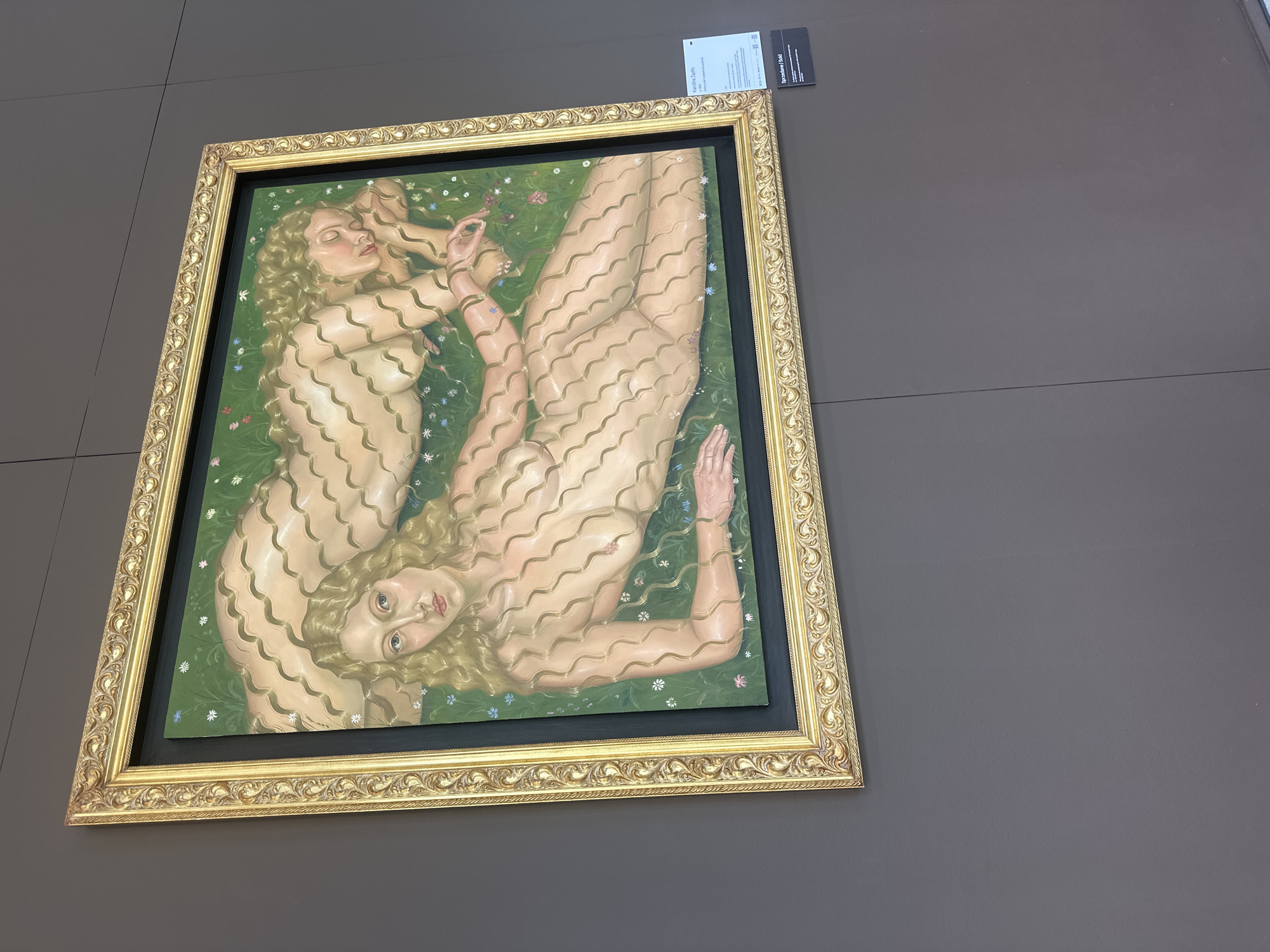 The image shows a painting titled "Karolina Zapłata" by Karolina Zapłata, dated 1990. The painting depicts two nude female figures with blonde hair, lying on a green, flower-strewn ground. The figures' bodies are adorned with wavy, ribbon-like patterns. The painting is framed in an ornate gold frame. To the right of the painting, a small white card with text in Polish and English provides information about the artwork and indicates that it is "Sprzedane / Sold." Below this card is a black label with text. The artwork is displayed on a dark gray wall, likely in an art gallery or exhibition space. Based on the labels and the style of presentation, this appears to be an exhibition in Warsaw, Poland.