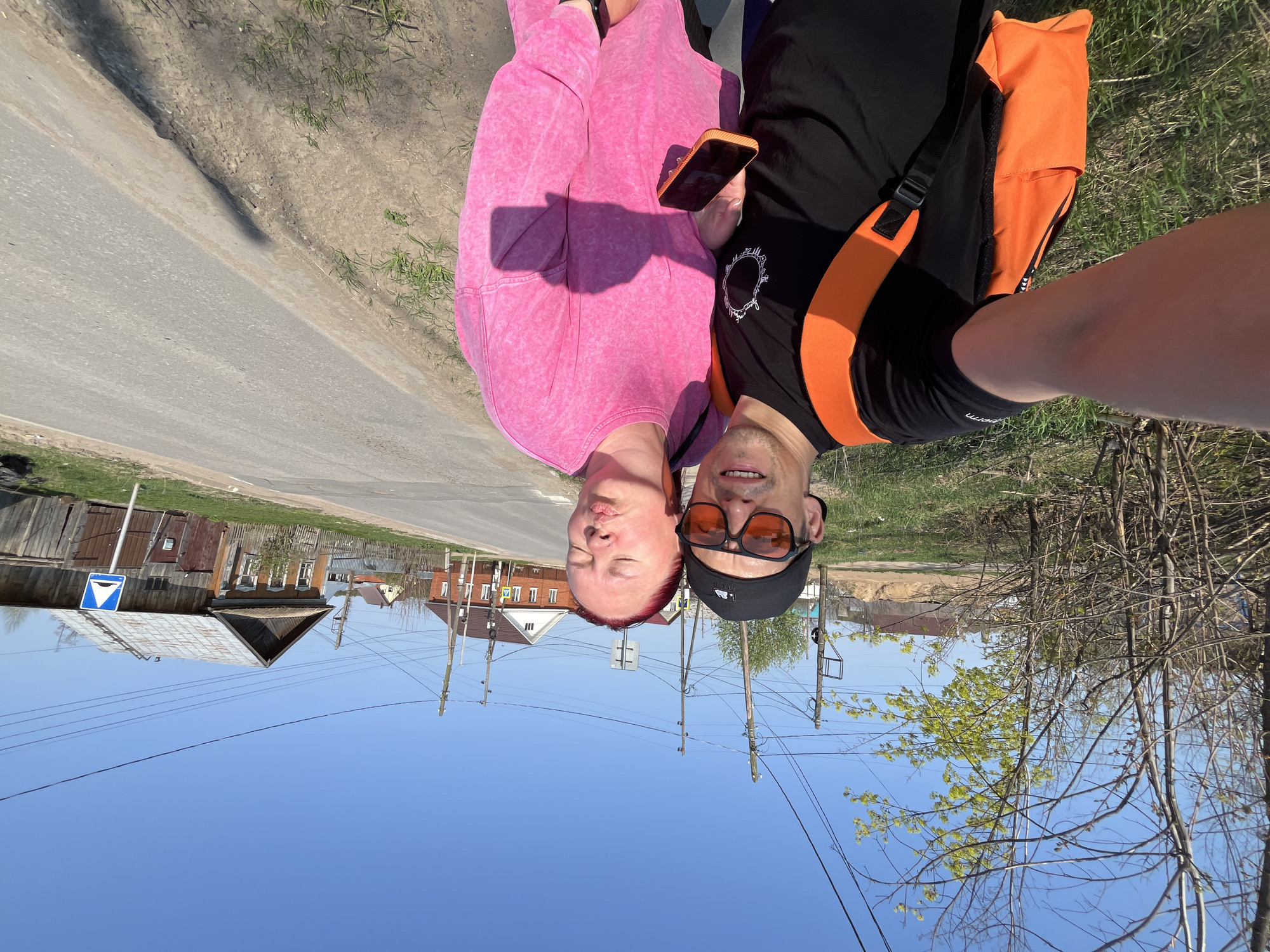 A selfie shows two people, a woman with closed eyes and a pink sweatsuit, and a man with sunglasses, a black hat, and a black shirt with orange straps, seemingly standing on a street in Chaikovsky, Russia. The image shows a typical Russian street view in the background. The sky is blue, and there are buildings, utility poles, and vegetation lining the street. The woman is holding an orange phone. The image is taken from a low angle, pointing upward.