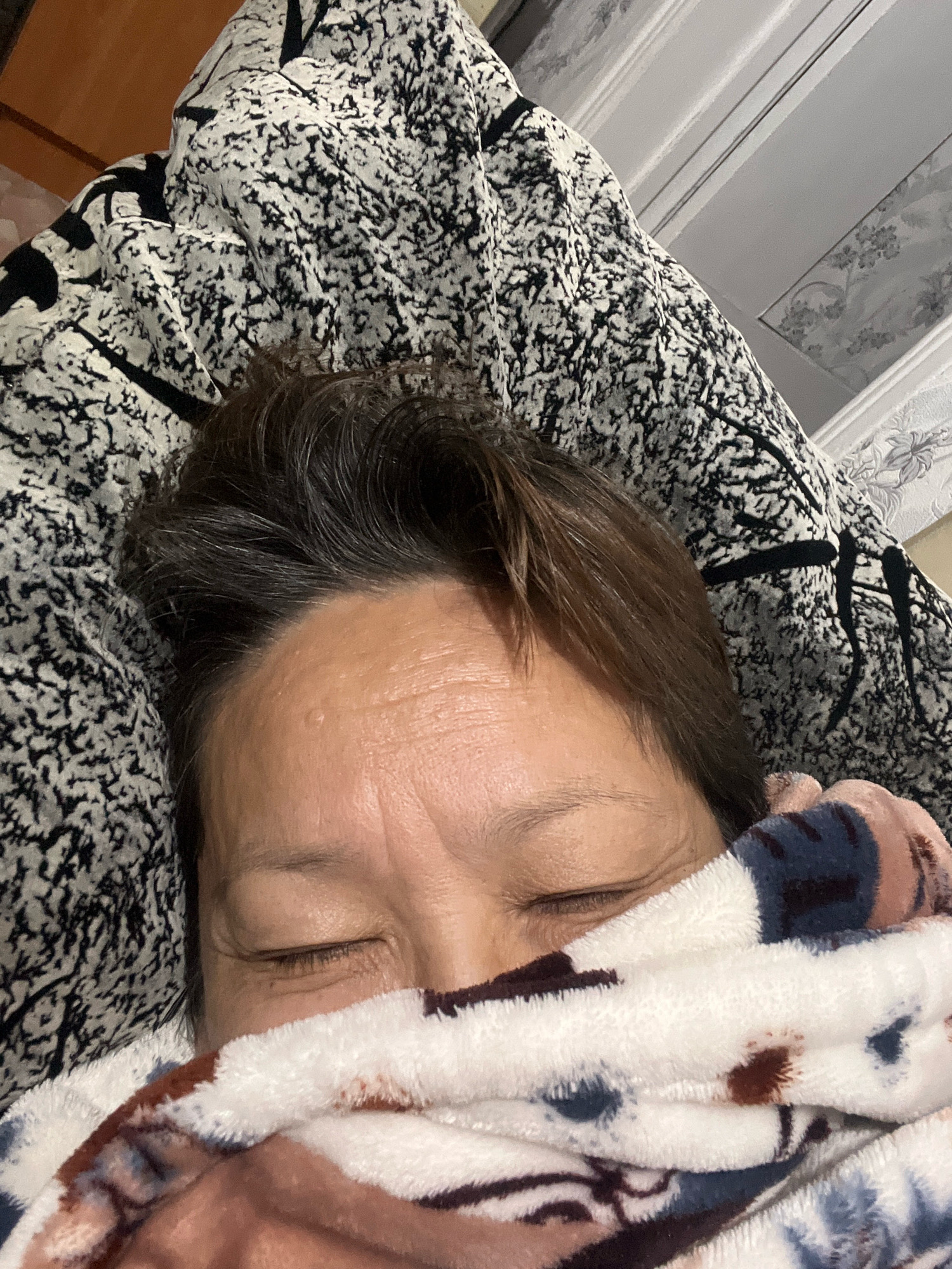 A person is lying down, partially covered by a patterned blanket. Their eyes are closed, and their forehead is visible, showing some wrinkles. The background consists of a bed with a patterned comforter and what appears to be a headboard. The scene suggests a relaxed or sleeping posture. The location is in the city of Тимирязев ауылдық округі, Kazakhstan.