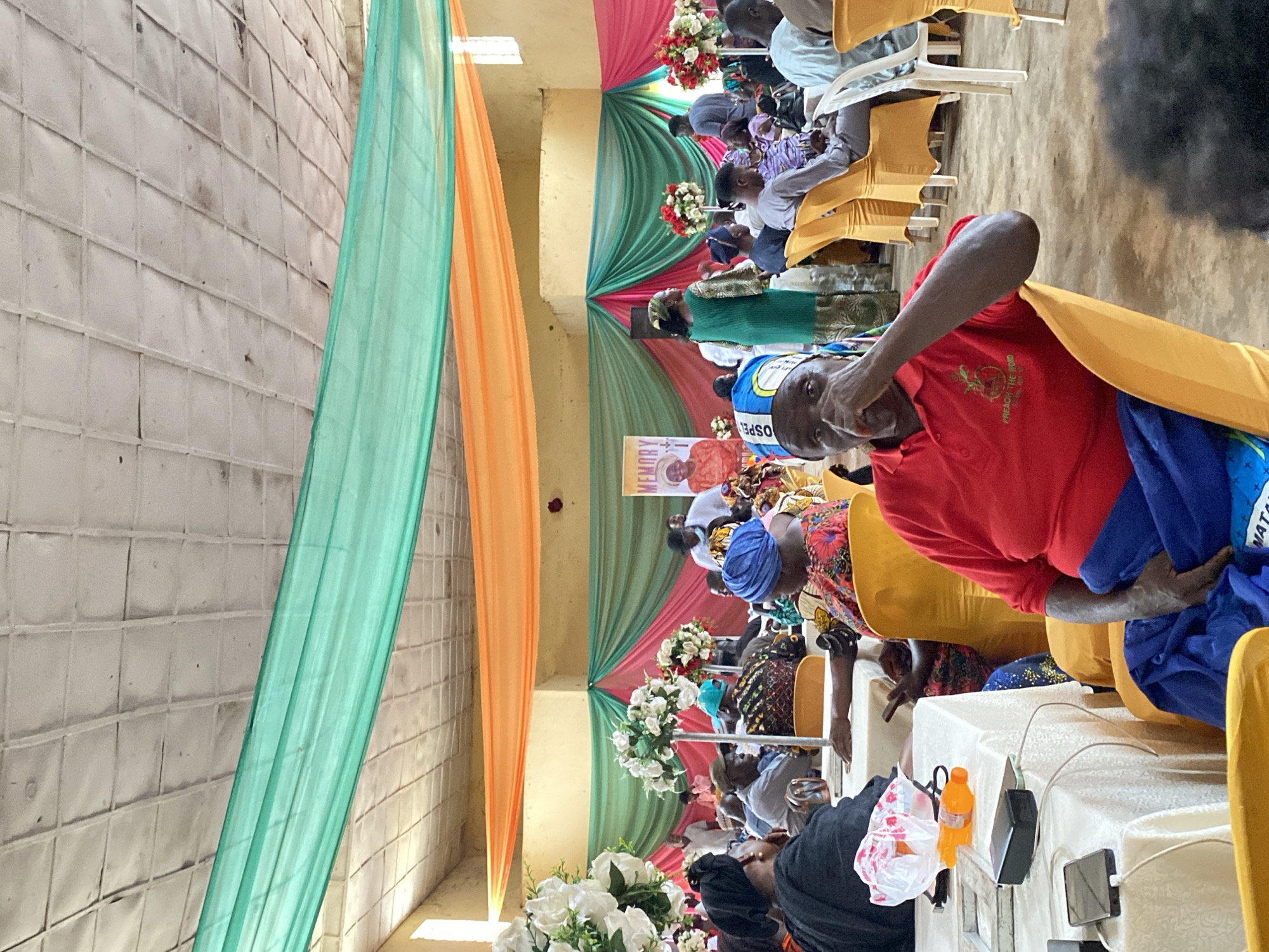 The image depicts an indoor gathering with people seated at tables, suggesting an event or ceremony. The room is decorated with draped fabric in vibrant colors of green, orange, and pink, which are also present in floral arrangements on tall stands. A banner with the word "MEMORY" and a portrait of a woman is visible in the background. The ceiling appears to be made of textured tiles. Several individuals are the focus of the foreground, including a man in a red shirt and blue wrapper looking towards the camera with his hand near his face, and other attendees dressed in traditional attire. There are objects on the tables, such as plastic bags, a bottle, and electronic devices like phones and a power bank connected to charging cables. The overall atmosphere suggests a communal event in Kuru, Nigeria.