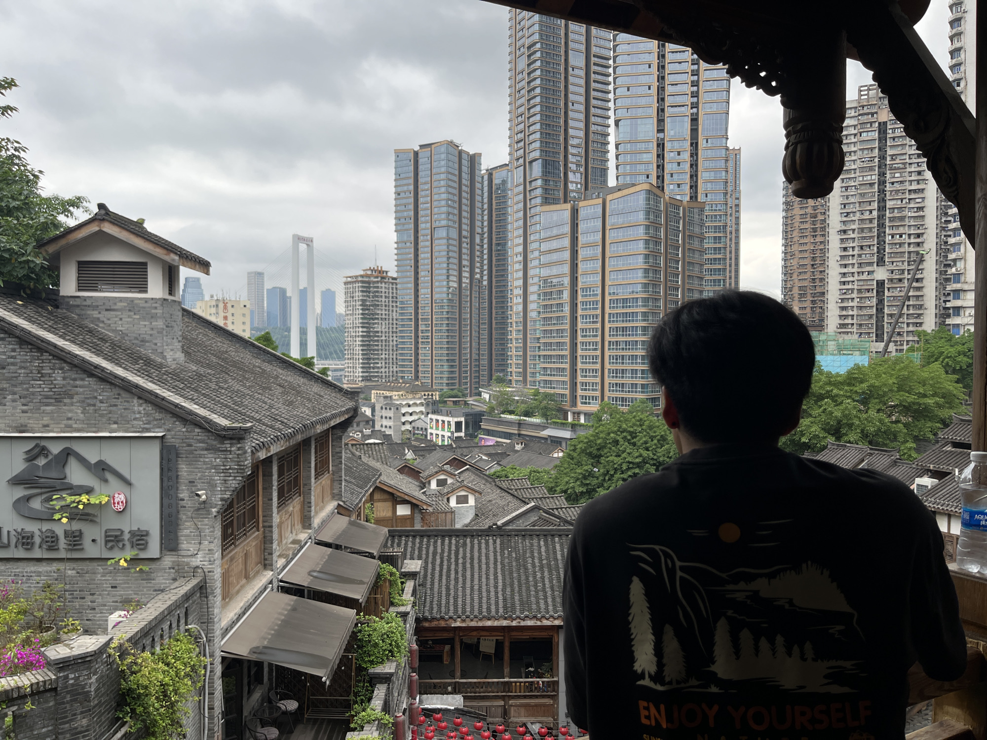 A medium shot presents a view of Yuzhong District, China, seen from an elevated vantage point. The back of a person wearing a dark t-shirt is partially visible in the lower right portion of the frame, obscuring part of the scene.
In the foreground and midground are traditional-style Chinese buildings with gray tiled roofs, some featuring covered walkways. Red lanterns are visible in a lower area. In the background, modern skyscrapers rise, partially obscured by a cloudy sky. A bridge can be seen in the distance, connecting parts of the cityscape. Vegetation is present, mixing with the urban landscape.
A sign with Chinese characters is visible on one of the buildings to the left. A water bottle rests on a ledge on the right side of the frame.