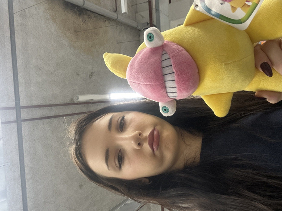 A close-up, eye-level shot captures a young woman holding a distinctive yellow plush toy. The image appears to be taken as a selfie, with the woman looking directly at the camera with a neutral expression.
The woman occupies the lower portion of the frame. She has long, dark brown hair that falls over her shoulders and part of her chest. Her skin is fair, and she has light makeup, including defined eyebrows, subtle eyeliner, and dark pink or reddish-brown lipstick. She is wearing a dark-colored top, possibly black or dark blue, visible below her chin.
To the right and slightly above the woman's face, a bright yellow plush toy is held up by her hand. The toy features a large, cartoonish pink mouth with prominent white teeth that are outlined in black, giving it a wide, toothy grin. Two large, googly eyes with green irises and black pupils extend from the top and bottom of the pink mouth. The toy also has small, yellow fin-like protrusions on its body. A white tag with colorful graphics (including hints of green, orange, and blue) is visible attached to the top of the toy, near the woman's hand. The woman's hand is visible on the right, holding the yellow toy, and her fingernails are painted with a dark red or maroon polish.
The background is an indoor setting with an industrial or utilitarian appearance. It consists of a grey, concrete-like wall or ceiling with visible pipes and light fixtures. There are horizontal dark lines, possibly support beams or conduits, running across the concrete surface. Patches of darker discoloration or stains are visible on the grey surface in the upper left portion of the image. The lighting appears to be artificial, likely from overhead fixtures, casting a somewhat cool tone on the scene.