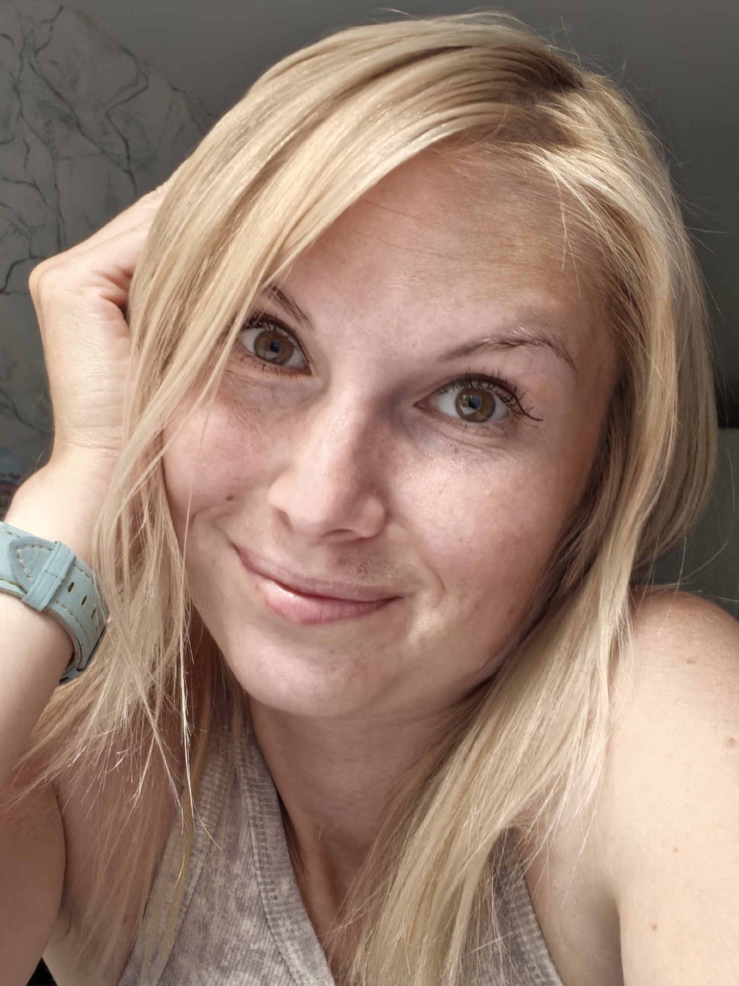 A close-up, head-and-shoulders selfie of a Caucasian woman with blonde hair, light skin, and brown eyes is shown. She is wearing a gray tank top and a light blue wristwatch on her left wrist. Her left hand is resting on her head. The background is blurred and appears to be a wall with a marble-like pattern. There is no information about the location in Sterlitamak, Russia.