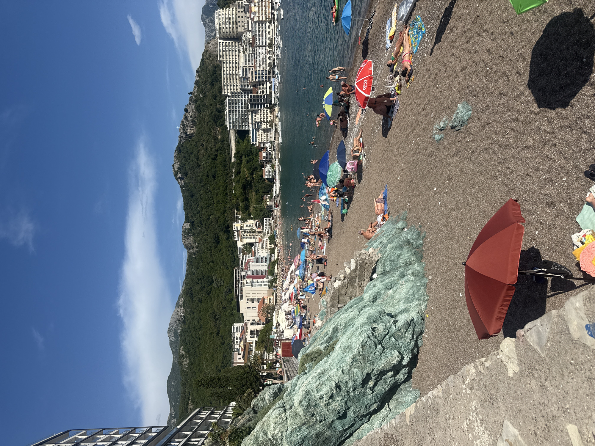 The image captures a bustling beach scene in Rafailovići, Montenegro, on a sunny day. The foreground features a large, reddish-brown umbrella, casting a shadow on the pebbly beach where several people are sunbathing and relaxing on towels and loungers. To the left, a large, textured green rock formation juts out, adding a natural element to the scene.
The mid-ground showcases a crowded beach with numerous umbrellas in various colors, including blue, green, and red, sheltering beachgoers. People are seen swimming in the calm, blue sea, wading near the shore, and enjoying the sun. The beach is lined with white buildings that ascend a steep, tree-covered mountainside, creating a dramatic backdrop. These buildings appear to be hotels or apartment complexes, characteristic of a popular coastal resort town.
In the background, the sea stretches out to the horizon, with a few boats visible. The sky is a clear, vibrant blue with a few wispy white clouds. The overall atmosphere is one of a lively summer day at a popular tourist destination. The image is taken from an elevated viewpoint, offering a panoramic perspective of the beach, town, and surrounding landscape.