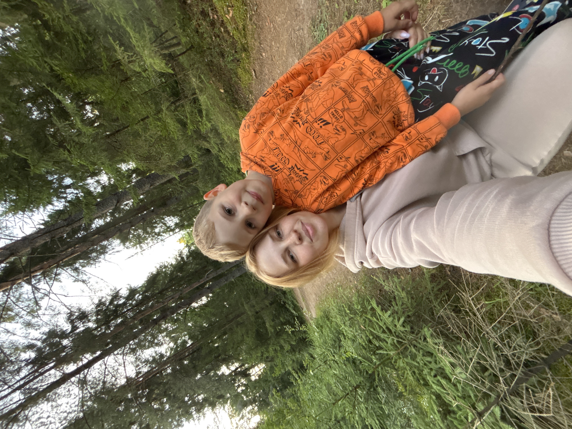 A young woman and a young boy are taking a selfie in a forest. The woman has blonde hair and is wearing a light pink hoodie. The boy has blonde hair and is wearing an orange hoodie with a dinosaur comic print and black pants with a colorful doodle pattern. They are standing on a dirt path, with dense green trees and foliage surrounding them. The sky is visible through the branches of the tall trees. The location is Anteplevo (Okhotinskoe S/P), Russia.