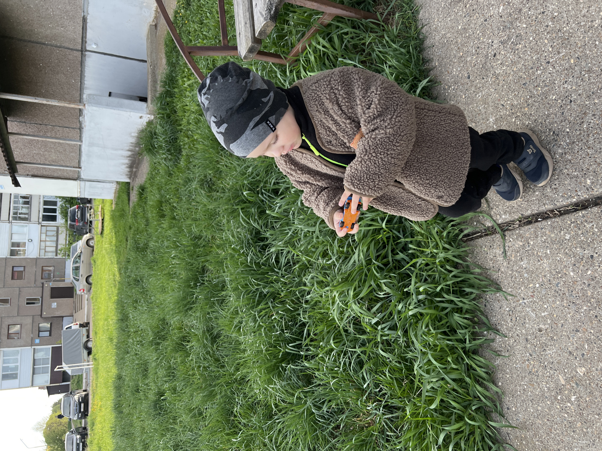 The image depicts a young child kneeling on a paved surface with grass on one side. The child is wearing a brown fleece jacket, black pants, and blue sneakers, and a camo beanie. The child is holding an orange toy. In the background is a residential building. Based on the location "Марковский, Russia", the scene appears to be an outdoor residential area in this city.