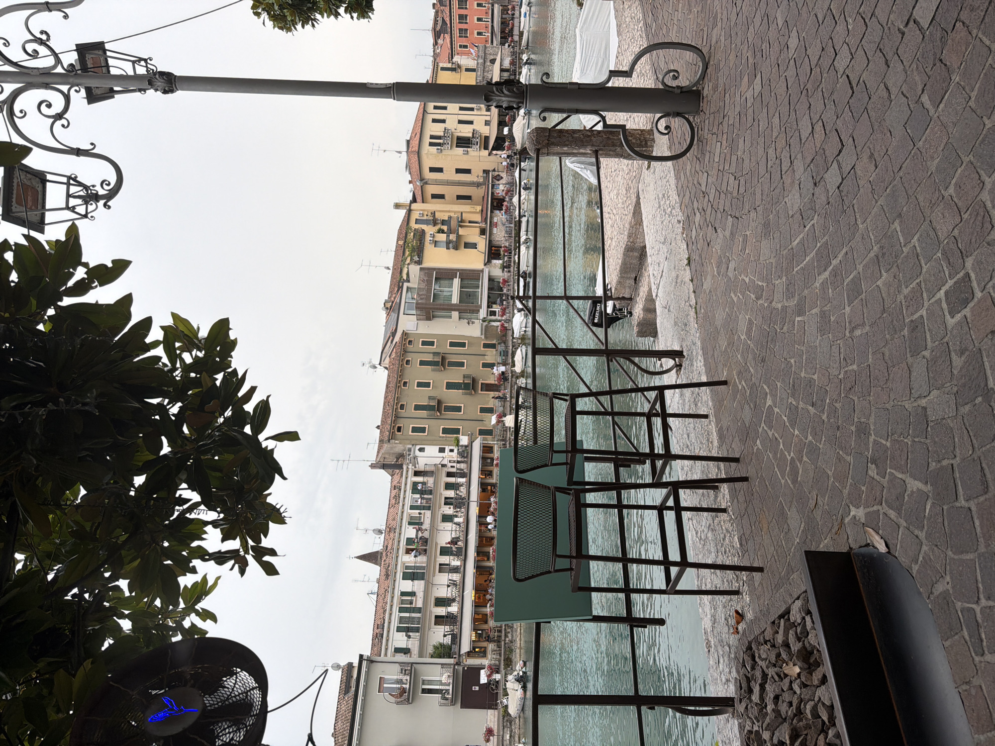 This image depicts a waterside promenade in Peschiera del Garda, Italy. In the foreground, two dark metal bar stools and a table with a green mesh surface are positioned near a railing overlooking a canal. The canal water is a greenish-blue color and appears calm. Several boats are docked on the opposite bank.
Behind the canal, a row of buildings with multi-story facades lines the waterfront. These buildings exhibit a variety of architectural styles and colors, ranging from pale yellow to cream and terracotta. Many have balconies with railing, and some have awnings over ground-floor windows, suggesting commercial establishments such as restaurants or cafes. People are visible on the balconies and sidewalks of these buildings.
The ground in the foreground is paved with cobblestones, arranged in a radial pattern. To the left of the stools, there is a dark metal planter filled with dark stones and a few fallen leaves. A large tree with dark green leaves partially obscures the upper left portion of the image, with a circular fan-like object mounted on a pole also visible. A decorative street lamp with multiple light fixtures stands to the right of the frame, its ornate metalwork a prominent feature. The sky is overcast.