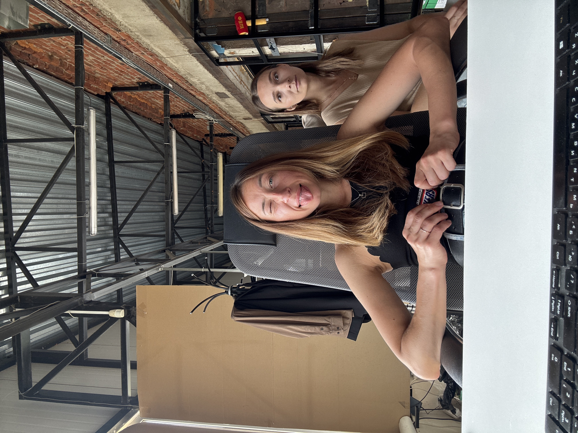 Here's a description of the image:
**Scene:** An interior space in what appears to be an office or studio environment, likely in Chaikovsky, Russia, based on the OCR text. The setting is characterized by a blend of industrial and contemporary design elements.
**People:** Two young women are present. The woman in the foreground is sitting in a chair and faces the camera. She is making a playful expression, sticking out her tongue and winking. She has long, light brown hair. She is wearing a black top and holding a watch. The second woman is sitting behind her and looking towards the camera. She has long, dark hair. She is wearing a beige top.
**Objects:** Visible objects include:
* A white desk with a keyboard.
* A chair.
* A folded brown jacket is hanging behind the chair.
* A corrugated metal ceiling structure.
* Industrial piping and fixtures.
* A cardboard panel.
**Actions/Interactions:** The primary action is the woman in the foreground posing for the camera. It's likely the two women are together in a work or social setting.
**Environment:** The space exhibits an industrial-chic aesthetic with exposed ceiling beams, corrugated metal, and brick. The lighting appears to be a mix of natural and artificial, with fluorescent or LED tube lights visible in the ceiling.