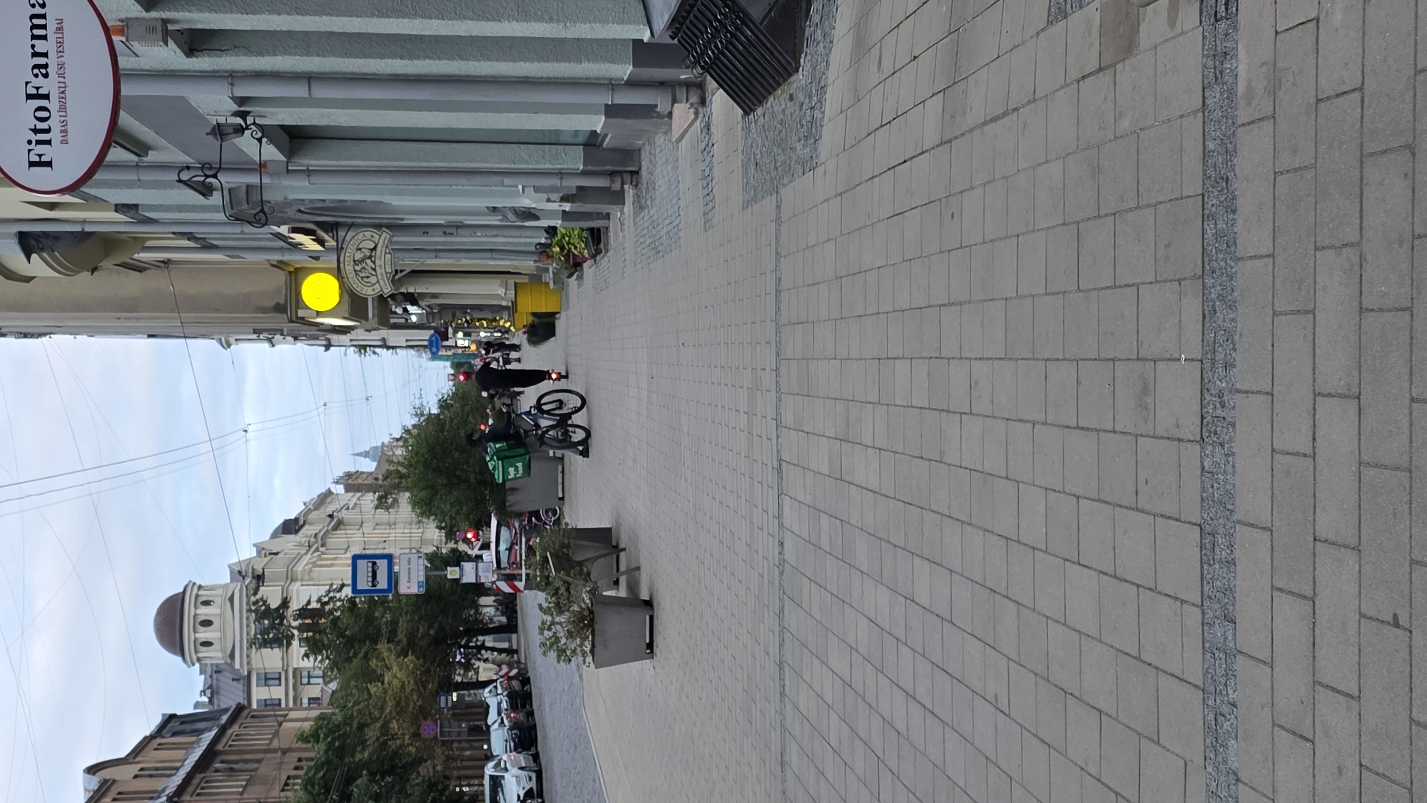 The image depicts a street scene in Riga, Latvia. A man is riding a scooter down a sidewalk paved with grey rectangular tiles. He is wearing dark clothing and a red light is visible on the back of his scooter. To his left, another person on a bicycle with a large green delivery bag is visible. The sidewalk is lined with large planters containing sparse green foliage. In the background, buildings with architectural details are visible, including a building with a dome-shaped roof and a sign that reads "FitoFarma". Traffic lights and street signs are also present. The sky is overcast.