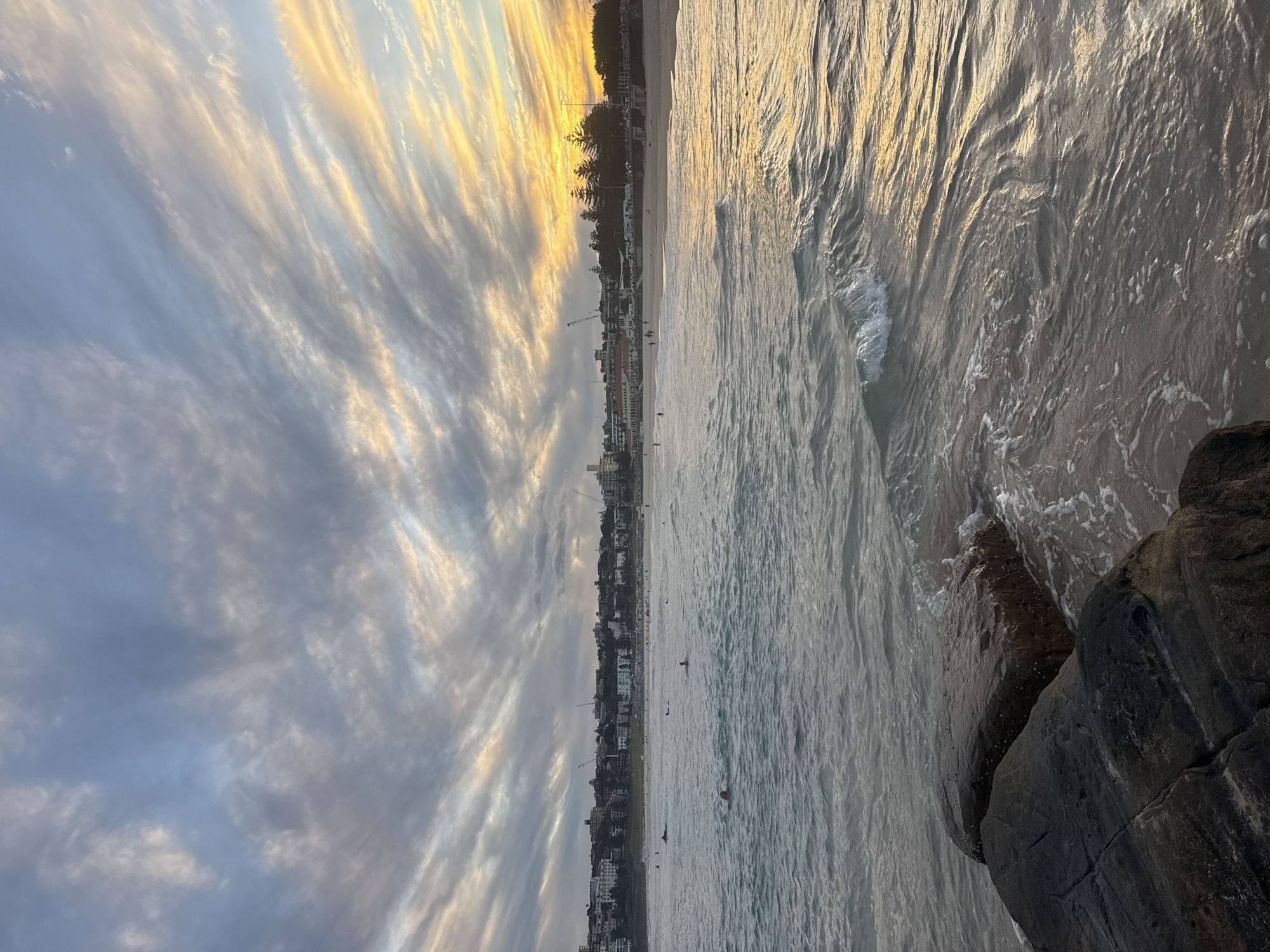 This is a photograph of Bondi Beach in Sydney, Australia, taken during sunset. The sky is filled with wispy clouds illuminated by the setting sun, creating a gradient of colors from soft blues to warm oranges. The ocean is calm with gentle waves lapping against the shore. In the foreground, dark, wet rocks are visible. In the middle ground, several people are swimming and surfing in the water. The background shows the Bondi Beach coastline with buildings and trees. Construction cranes are visible in the distance, suggesting ongoing development.