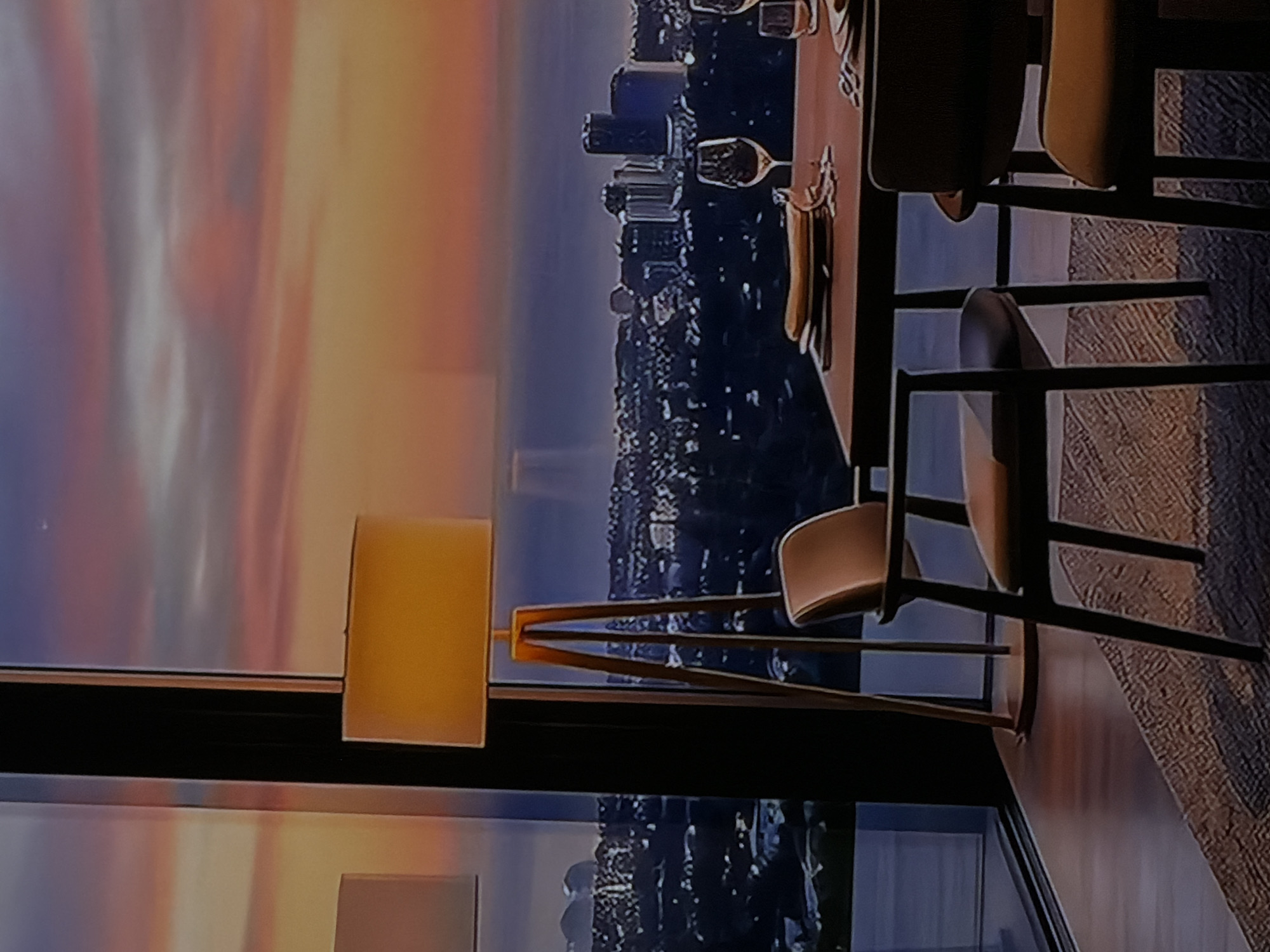 The image depicts a modern dining area within what appears to be a high-rise building, offering a panoramic view of a city at dusk or dawn. The scene is presented from an interior perspective, looking out through a large, floor-to-ceiling window.
In the foreground and midground, a contemporary dining set is arranged. A wooden rectangular table is visible on the right, set with multiple plates, cutlery, and wine glasses, suggesting preparation for a meal or a recently concluded one. Several modern dining chairs with dark frames and light-colored, possibly upholstered, seats are pulled up to the table. To the left of the dining table, a stylish floor lamp with a rectangular, light-colored shade and a dark, possibly wooden or metal, stand illuminates the interior space. The floor beneath the dining area has a textured finish, resembling a carpet or large rug.
Beyond the expansive window, a vast cityscape unfolds. Numerous buildings, ranging in height, form a dense urban landscape, with lights beginning to twinkle or still present from the night. The sky above the city displays vibrant hues of orange, red, purple, and blue, indicative of either a striking sunset or sunrise, creating a serene and picturesque backdrop. The weather appears clear and calm.
No people are visible in the image, and no specific activity is actively occurring, but the set table implies a social gathering or meal. There is no visible text in the image. The setting suggests a sophisticated and elevated living or dining experience in an urban environment, specifically located in Sofiivska Borshchahivka, Ukraine, according to the provided context.