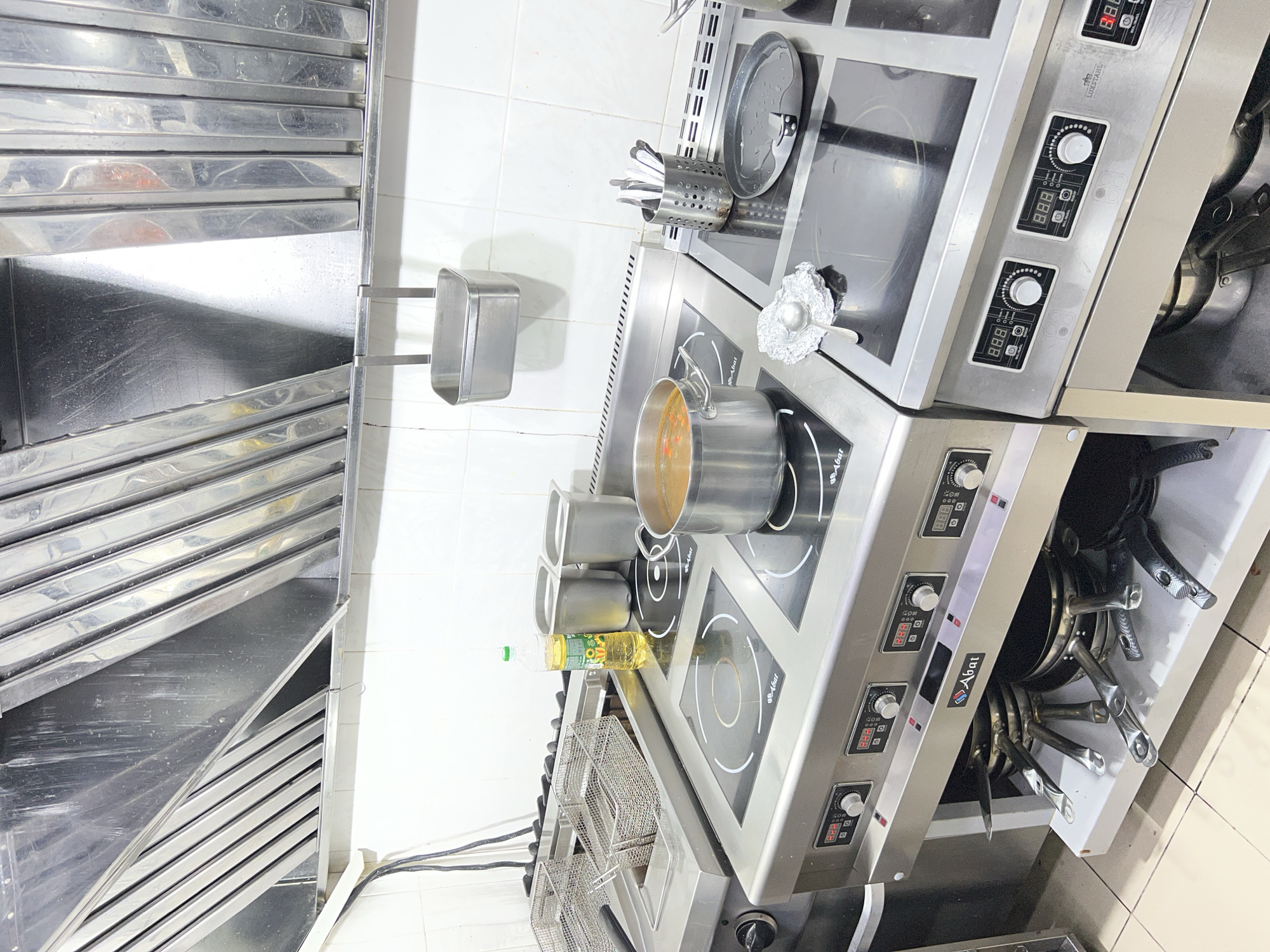 A commercial kitchen scene is depicted, focusing on a stainless steel cooking range with multiple induction burners. On the top surface, a large pot of soup simmers on one of the burners, alongside a bottle of cooking oil and two metallic containers. To the right, a utensil holder filled with various kitchen tools and a flat pan are visible. The kitchen is equipped with a commercial-grade exhaust hood overhead, a deep fryer on the left, and cabinets filled with pots and pans beneath the cooking surface. The environment is characterized by white tiled walls and a polished metallic aesthetic, suggesting a professional food preparation area. The image does not contain any people or overt actions, presenting a static view of the kitchen's setup. The location of Plastunivska, Russia, is provided as context for this scene.