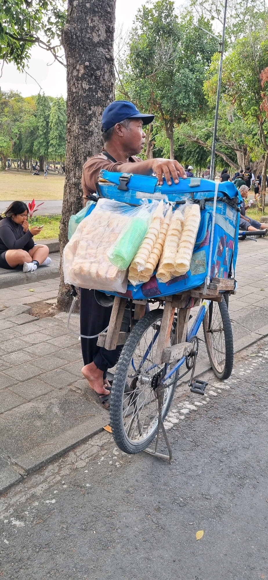 In Denpasar, Indonesia, a man in a blue cap and brown shirt stands next to a blue ice cream cart attached to a bicycle. The cart is loaded with ice cream cones and packaged snacks. To the left, a young woman in a black outfit sits on a curb, looking at her phone. The scene unfolds on a paved walkway lined with trees, suggesting a public park or recreational area.