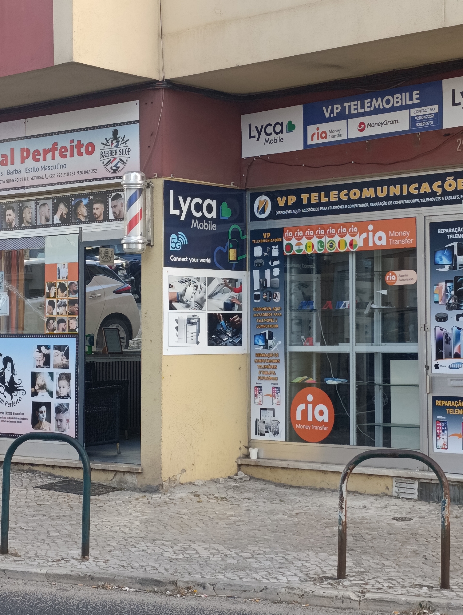 The image shows the exterior of a commercial establishment in Setúbal, Portugal. The storefront is divided into sections, with awnings and signage indicating various services. To the left, a barber shop displays images of haircuts and hairstyles. The shop's name, "al Perfeito," is prominently displayed, along with the words "Barber Shop." The address and phone number are also visible.
In the center of the image, a Lyca Mobile sign is visible, featuring the Lyca Mobile logo and the words "Connect your world." Below this, there are several smaller images showcasing mobile devices and accessories.
To the right, a sign for "V.P. TELECOMUNICAÇÕES" is displayed, along with logos for Lyca Mobile, Ria Money Transfer, and MoneyGram. This section appears to be a telecommunications store, offering accessories, computer repair, and phone services. Another sign for "REPARAÇÃO DE TELEMÓVEIS" (phone repair) is also visible.
The storefront is made of glass, allowing a view into the interior where shelves are stocked with products. A white car is parked partially visible in the background. The pavement in front of the store is paved with cobblestones, and a metal bicycle rack is positioned on the sidewalk. The overall scene depicts a busy commercial street.