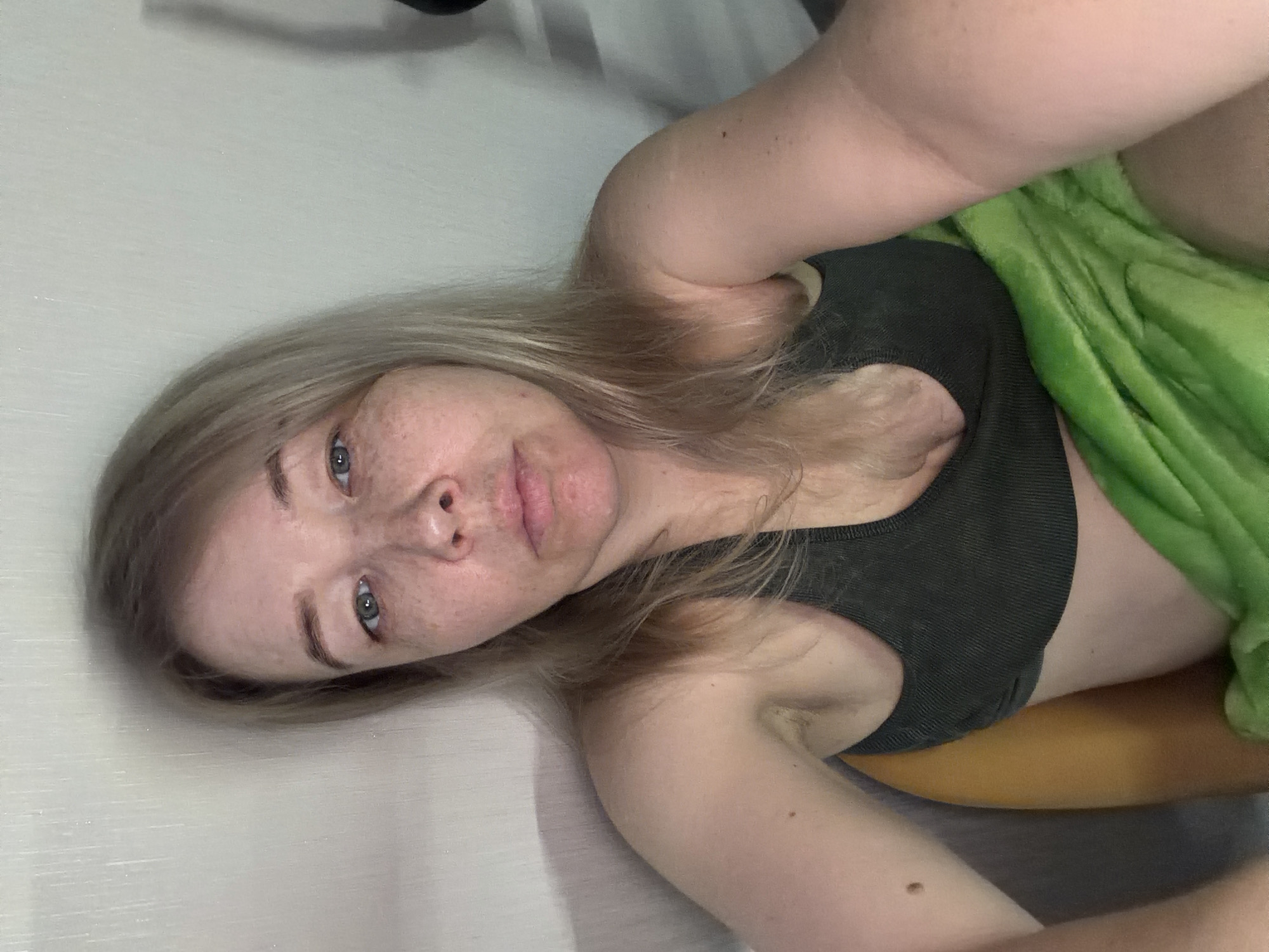 A woman is lying on a surface, possibly a bed, with a green blanket partially covering her torso. She is wearing a dark grey sports bra. The woman has blonde hair and visible freckles on her face. The image does not contain details to identify any location.