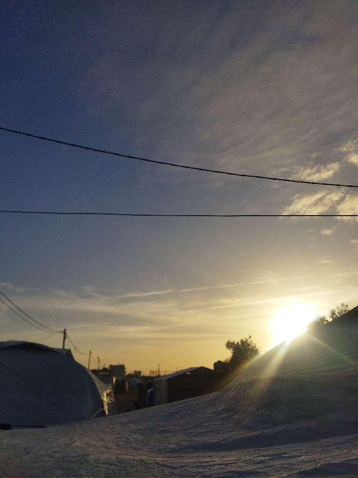 The image depicts an outdoor environment at either sunrise or sunset in Khan Yunis, Palestinian Territory. The sky transitions from deep blue at the top to a vibrant yellow-orange at the horizon on the right, featuring scattered wispy clouds. Two prominent, dark horizontal utility lines traverse the upper mid-section of the frame. The sun, a bright source of light, is positioned low on the right horizon, partially obscured by an elevated dark landform and emitting distinct light streaks. The foreground consists of undulating, light-colored ground, consistent with sand or earth, displaying texture from low-angle illumination. On the far left, a large, rounded, light-toned tent or temporary shelter is partially visible. On the right, a dark, vegetated slope ascends, partially concealing the sun. In the midground, a collection of indistinct, low-profile structures, likely additional tents or makeshift buildings, along with utility poles, are silhouetted against the bright horizon. No individuals are present, and no actions or interactions are observed.