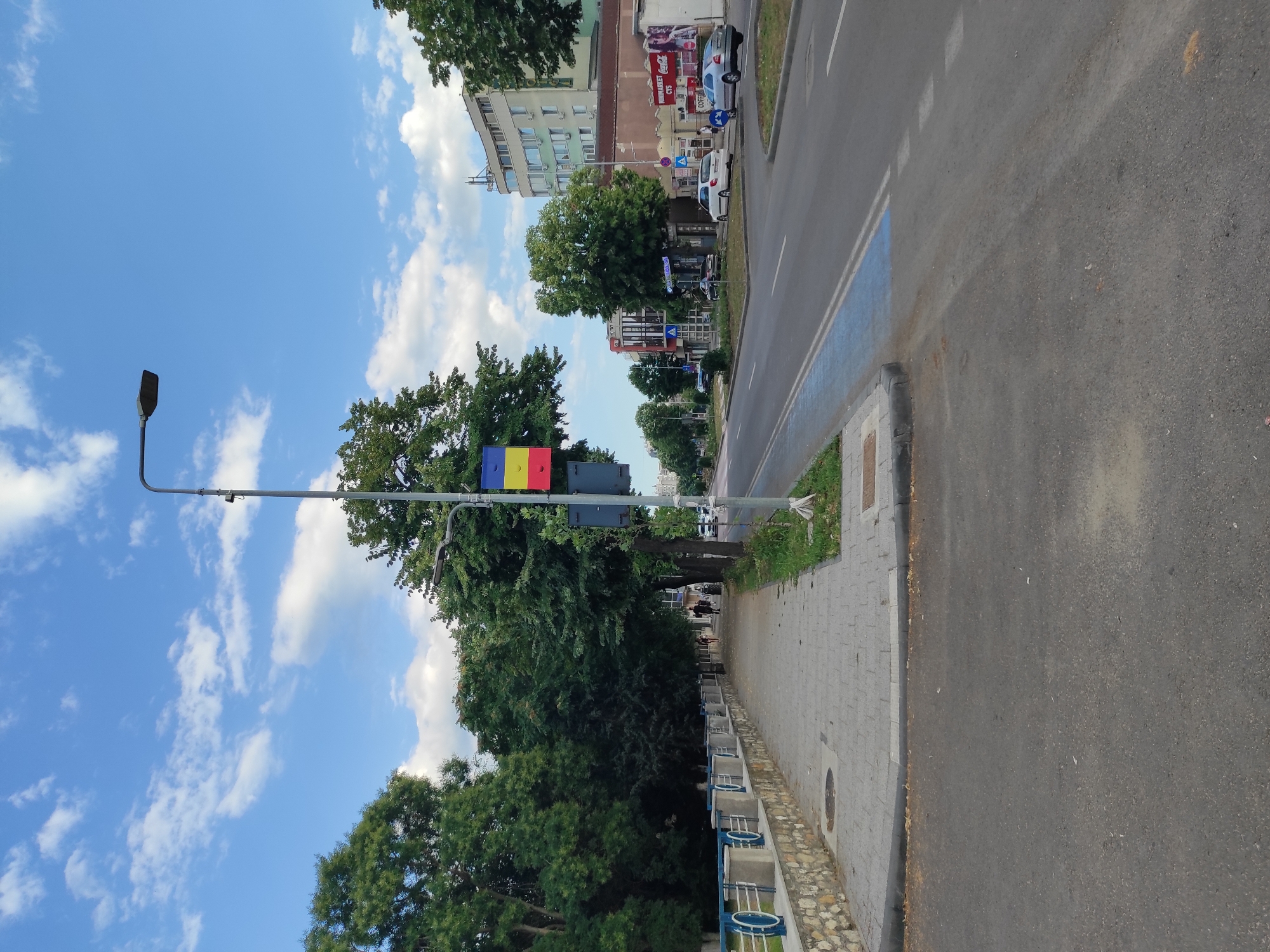 The image depicts a street scene in Constanța, Romania, likely during daytime. The foreground is dominated by an asphalt road with visible lane markings, including a blue painted section that may indicate a bike lane or a special zone. To the left of the road, a paved sidewalk runs alongside a stone and concrete wall with blue railing elements. Lush green trees line the sidewalk, providing shade.
Further down the street, several buildings of varying architectural styles are visible, including a multi-story building with a curved facade. A street light stands prominently near the sidewalk, displaying three colored rectangular flags (likely the Romanian flag: blue, yellow, and red) attached to it. A few cars are parked or driving on the street, and a couple of pedestrians are walking on the sidewalk. The sky is partly cloudy with a bright blue sky. A Coca-Cola advertisement is visible on one of the buildings. The overall impression is that of a typical urban street in Romania, with elements of nature and commerce coexisting.